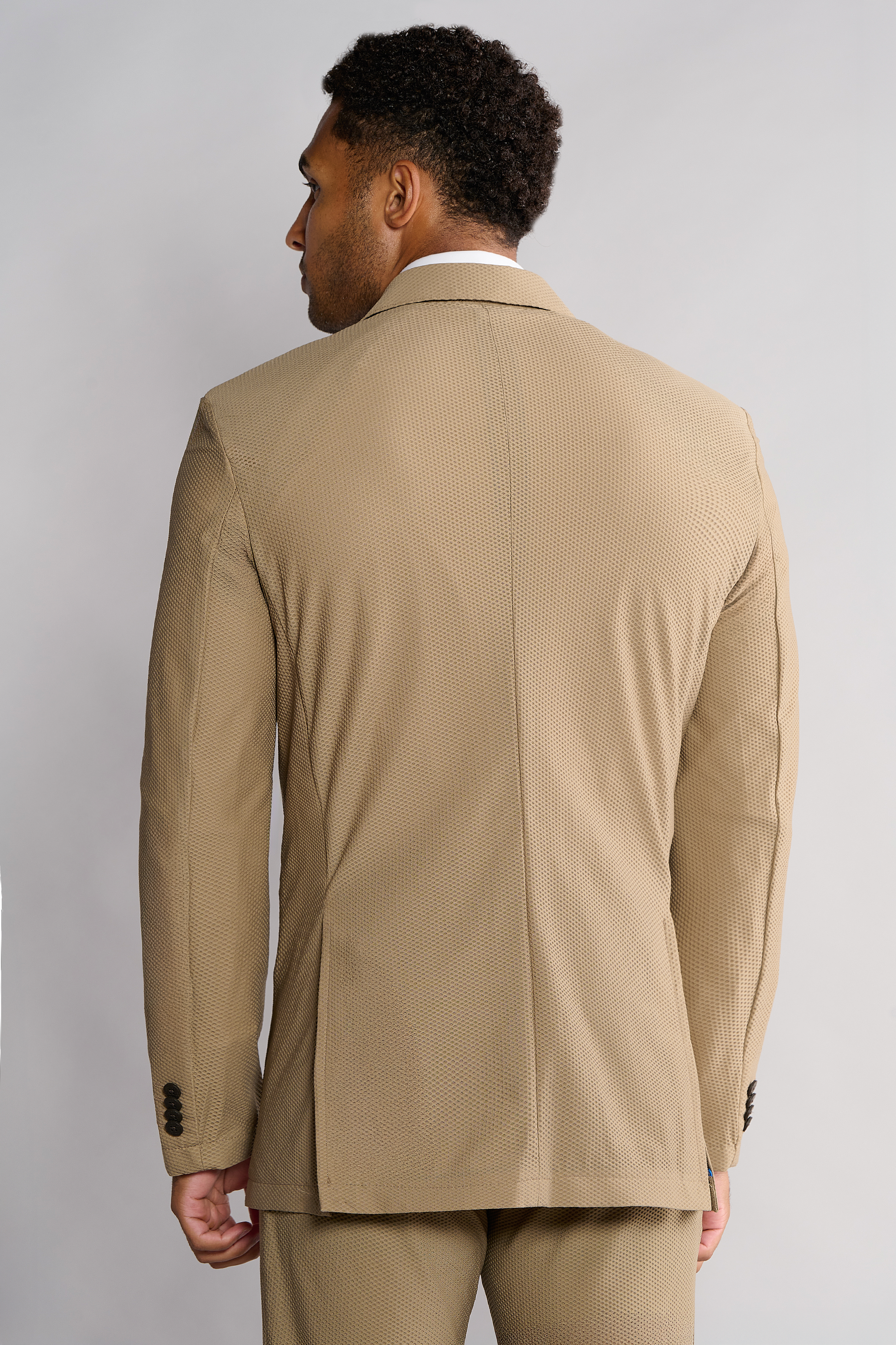 A man with short, curly hair faces away, wearing the Twillory AIR Blazer // Khaki. The blazer features 360° airflow fabric, a subtle pattern, two-button cuffs, and a center back seam against a light gray background.