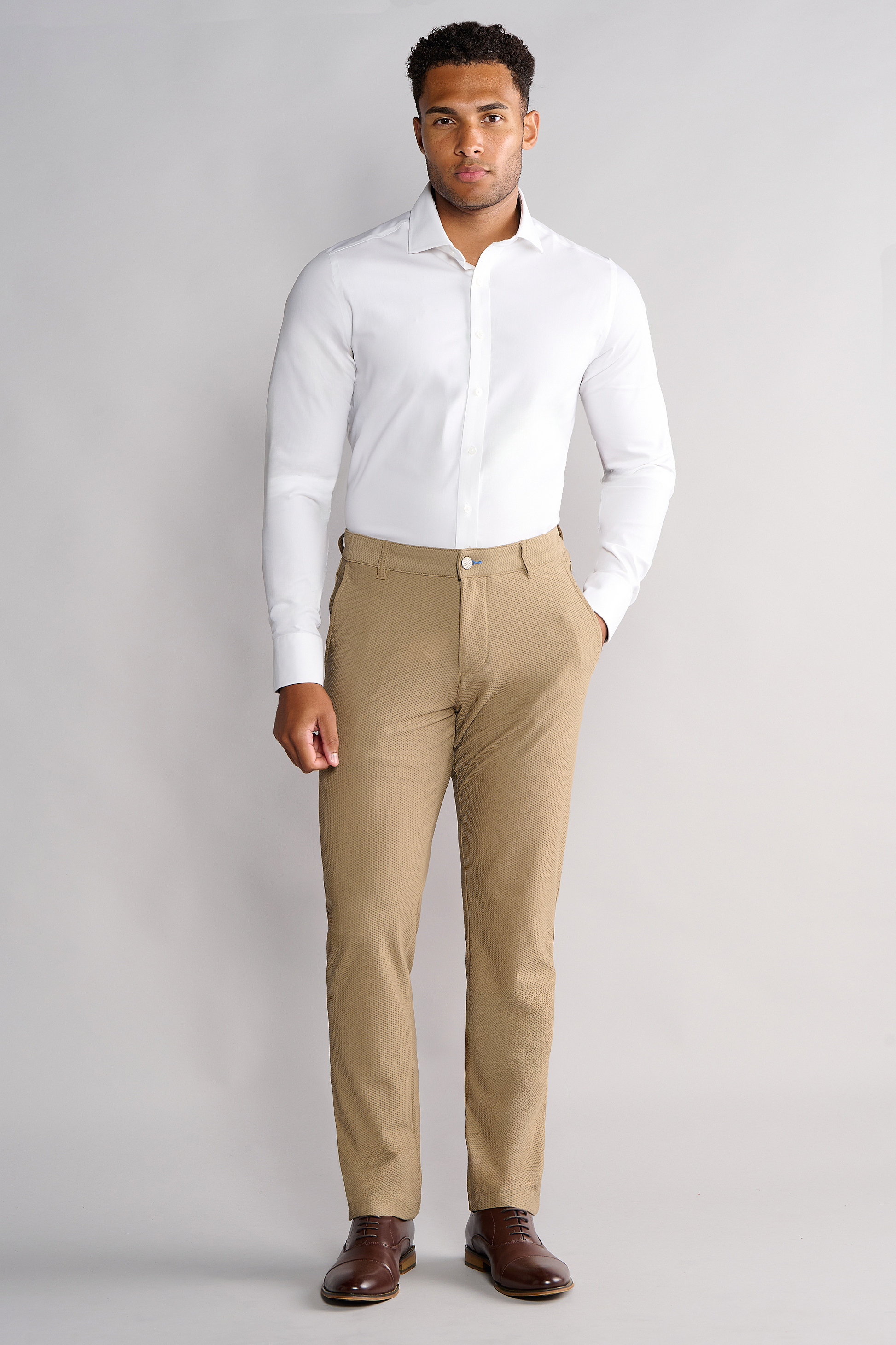 A man stands against a light gray background wearing Twillory AIR Pants in Khaki, a fitted white dress shirt, and brown dress shoes. He has one hand in his pocket and looks directly at the camera with a neutral expression.