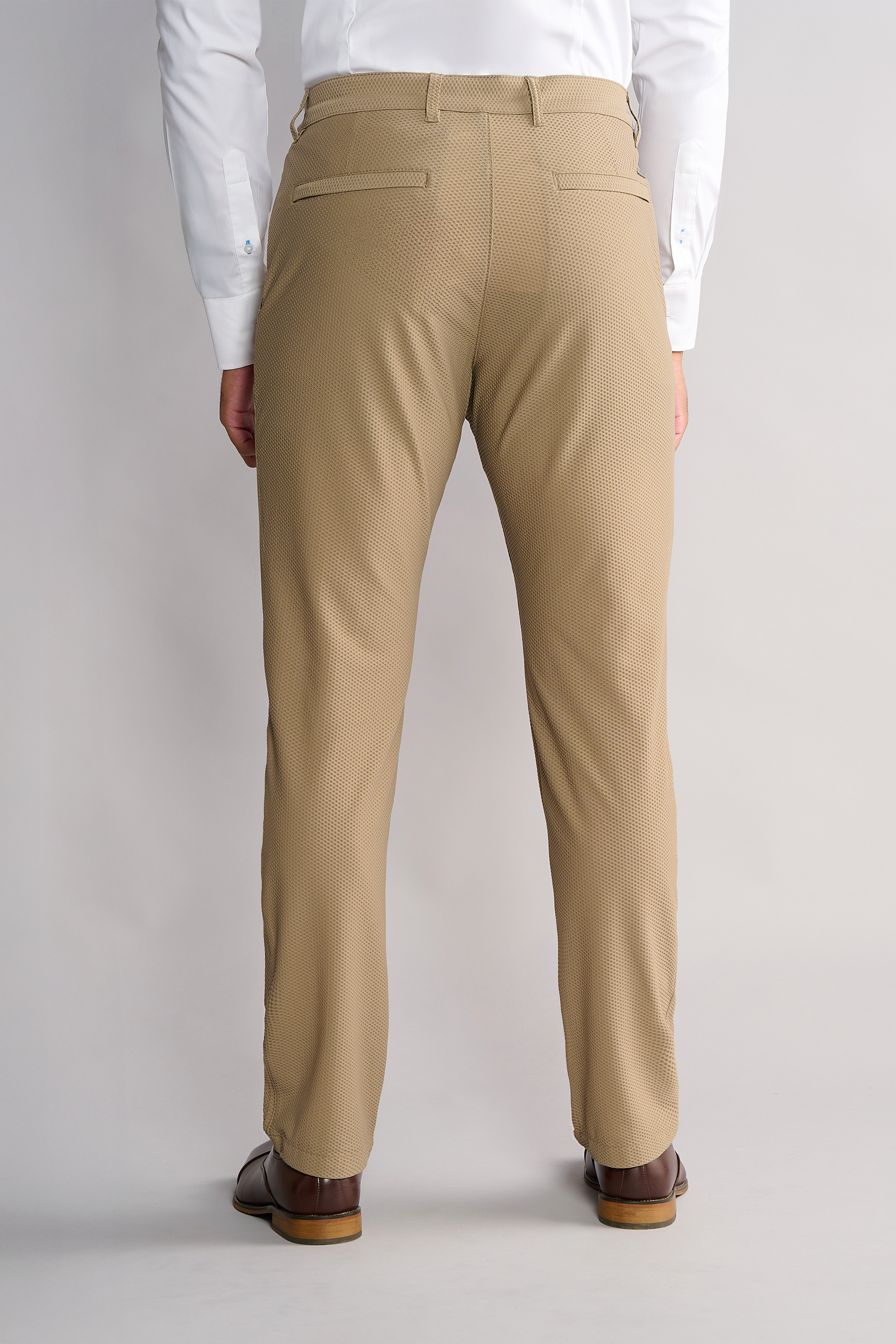 A person stands facing away, wearing Twillory AIR Pants // Khaki, a white button-down shirt, and brown dress shoes. The pants feature two back pockets with buttoned flaps. The background is plain and light-colored.