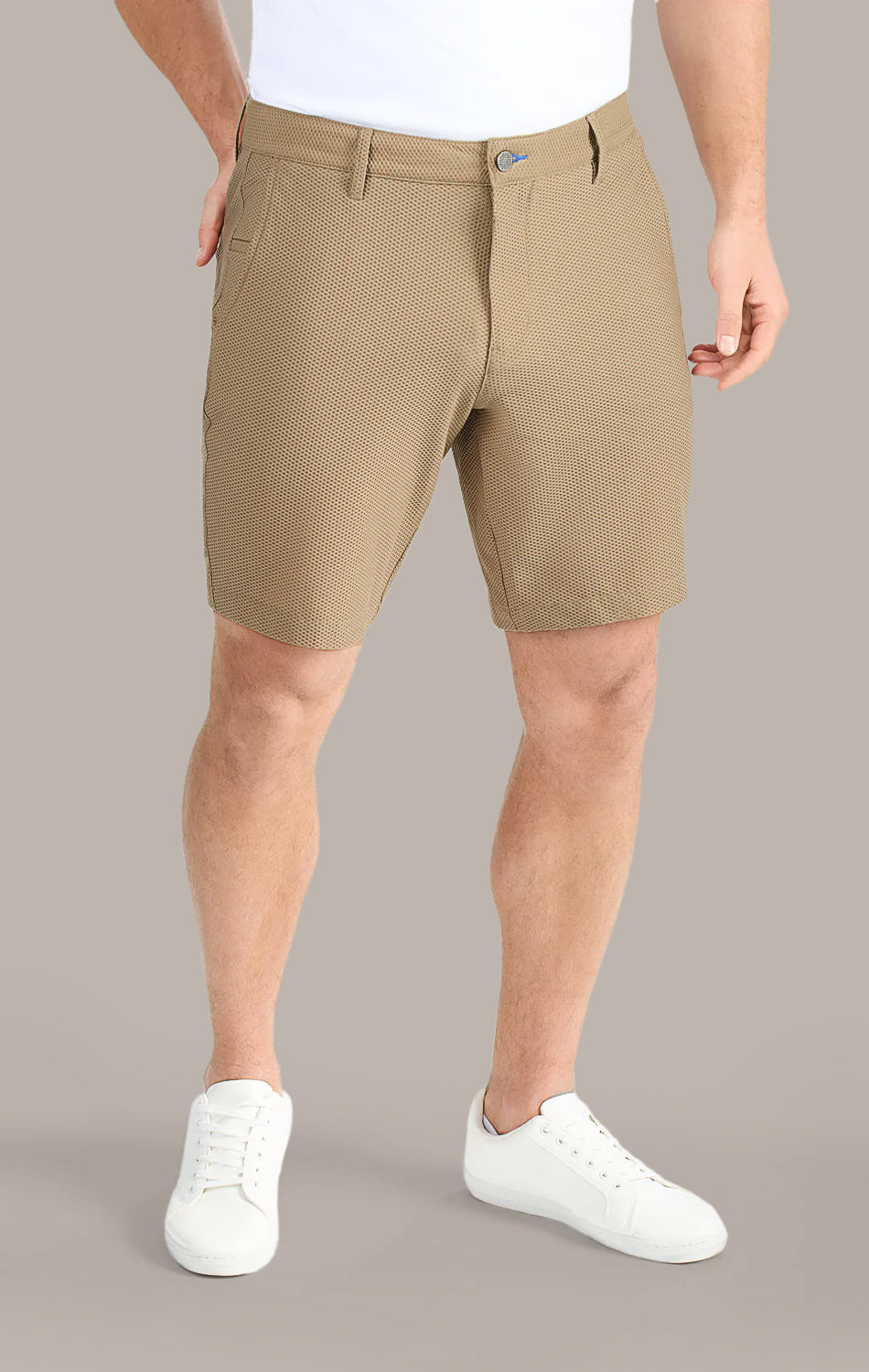 A man models Twillory AIR Shorts—light brown, 4-way stretch, sweat-wicking—paired with a white tucked-in shirt and white sneakers. His torso and legs are shown against a plain background, one hand in his pocket.