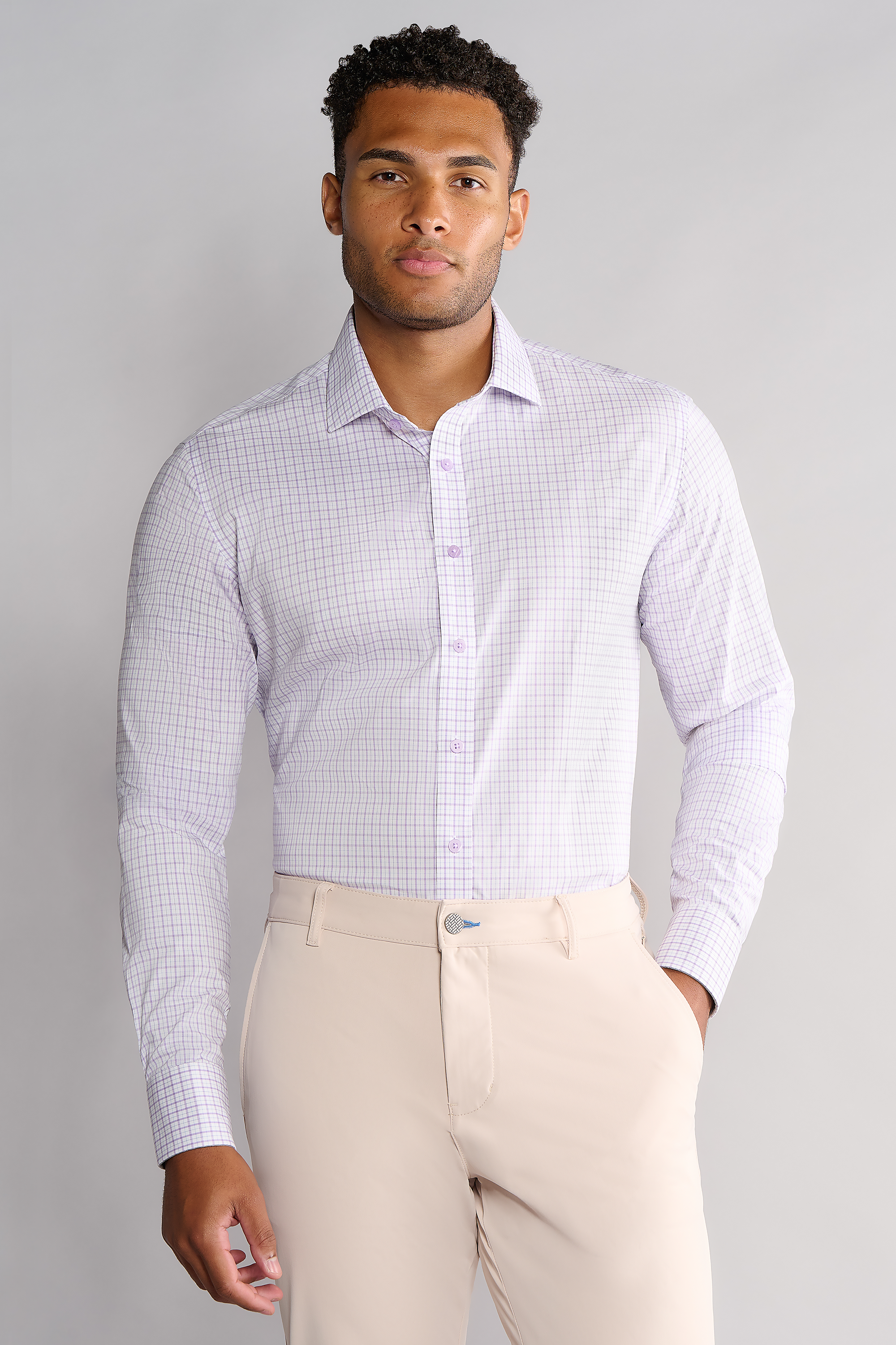 A man with short curly hair and a neutral expression wears the Twillory Leader Windowpane // Lavender performance shirt tucked into beige dress pants. He stands facing forward against a plain light gray background with soft, even lighting.