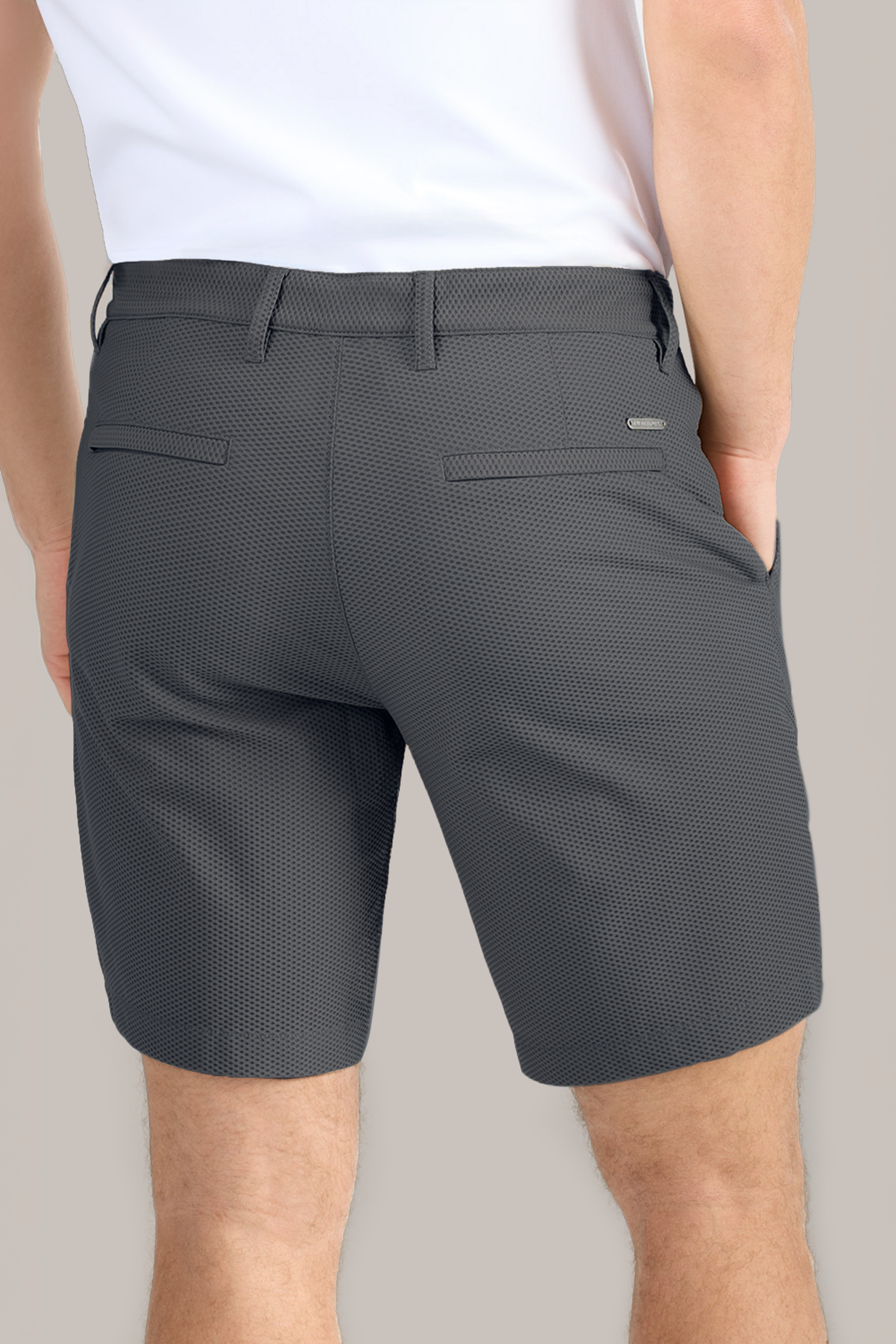 A person wearing a white shirt and Twillory AIR Shorts in textured dark gray 4-way stretch stands with one hand in their pocket. The back view highlights belt loops, two welt pockets, and a plain light background.