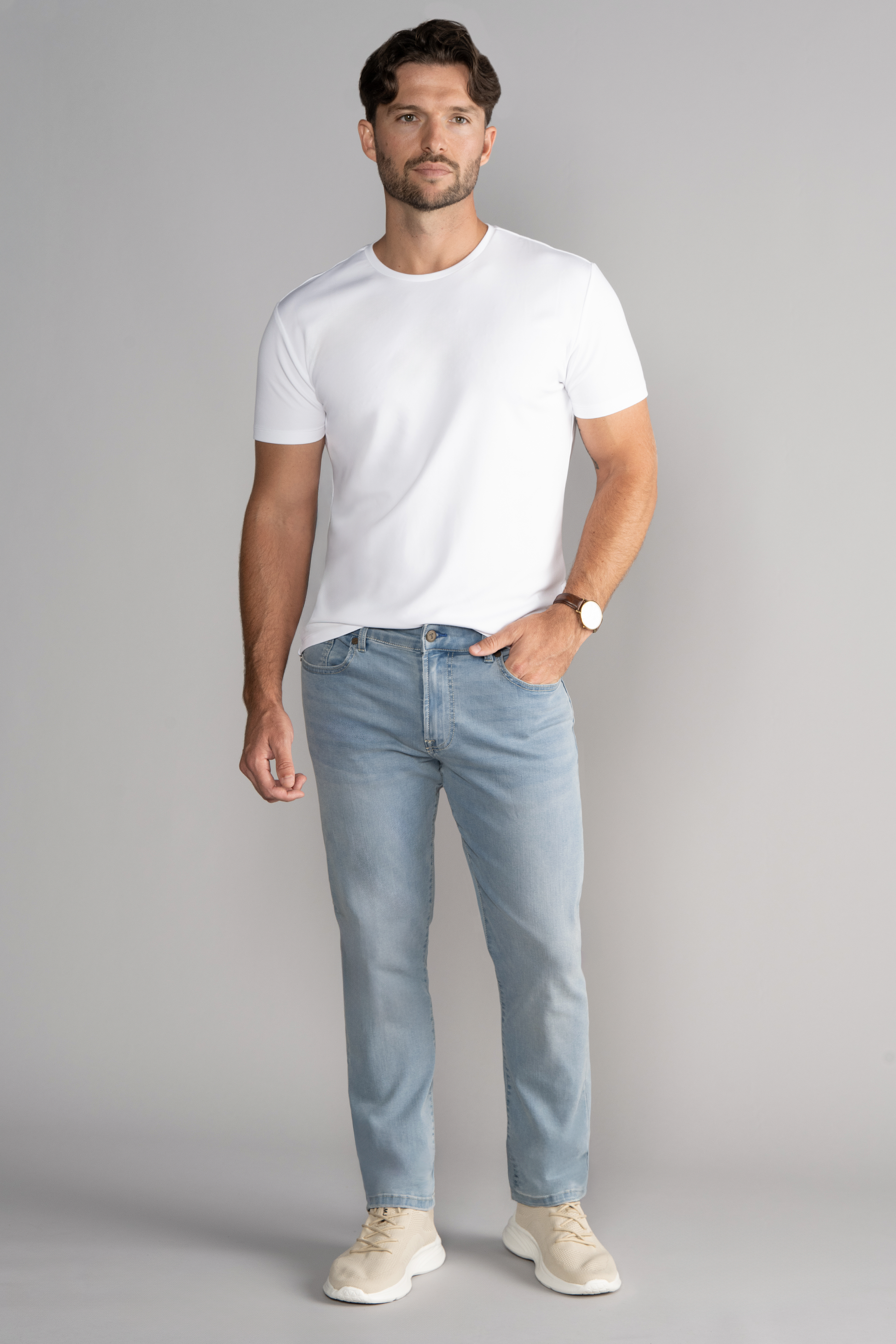 A man with short brown hair and a beard stands against a gray background, wearing a fitted white t-shirt, beige sneakers, brown wristwatch, and Twillory Performance Denim // LIGHT WASH BLUE jeans with a tapered fit—a hand in his pocket, expression neutral.