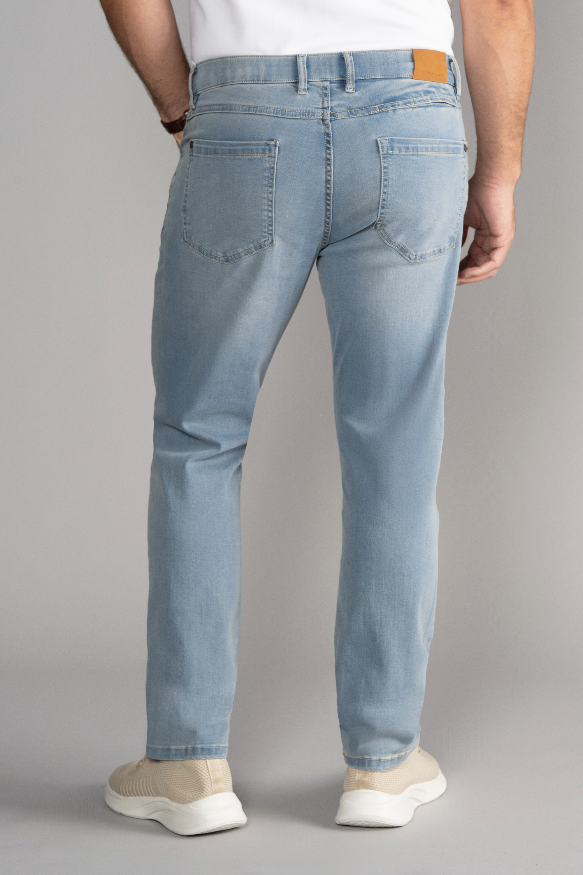 A person is shown from the back wearing Twillory Performance Denim // LIGHT WASH BLUE jeans with a tapered fit, two back pockets, a brown waistband patch, beige sneakers, and a white top against a plain gray background.