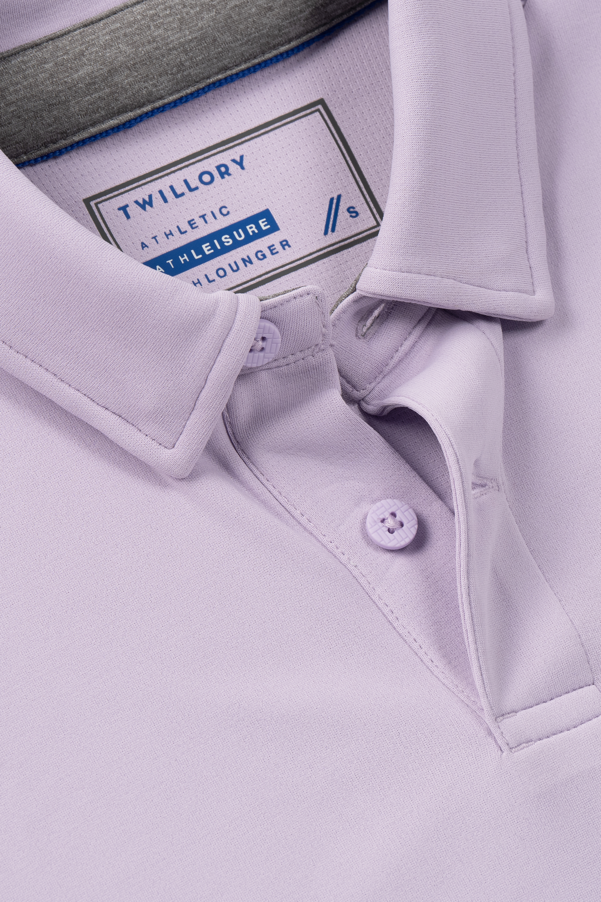 Close-up of the Twillory Short Sleeve Polo Solid in Lilac, featuring a collar, two buttons, and a visible neck tag reading "Twillory" in navy blue and gray. Moisture-wicking performance design. Style: Athleisure Lounger, Size: M/S.