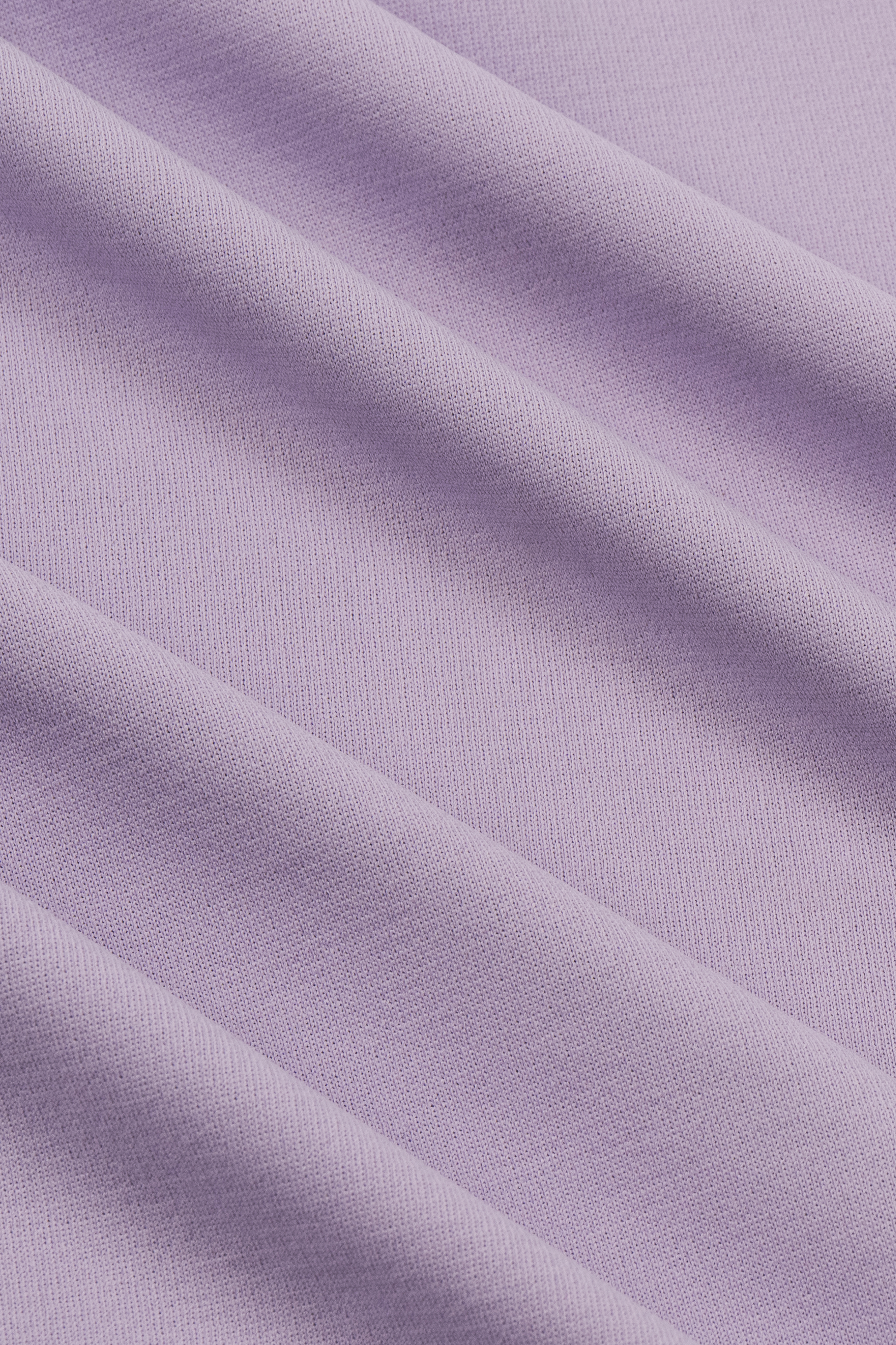 Close-up of lilac fabric from the Twillory Short Sleeve Polo Solid, showing soft diagonal folds. Its smooth, matte moisture-wicking texture and subtle shadows give the shirt a calm and elegant look.