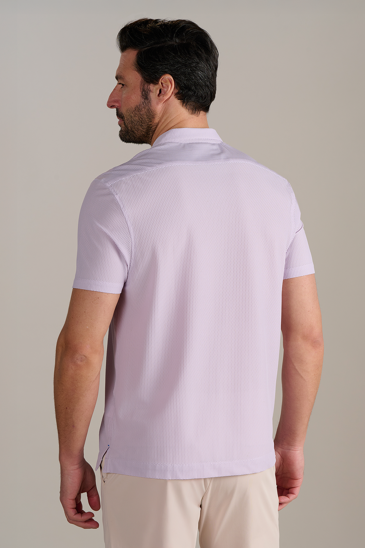 A man with short dark hair and facial hair stands with his back to the camera, wearing Twillory's AIR Resort Shirt Solid in Lilac, which is machine washable, paired with beige pants against a plain, light background.