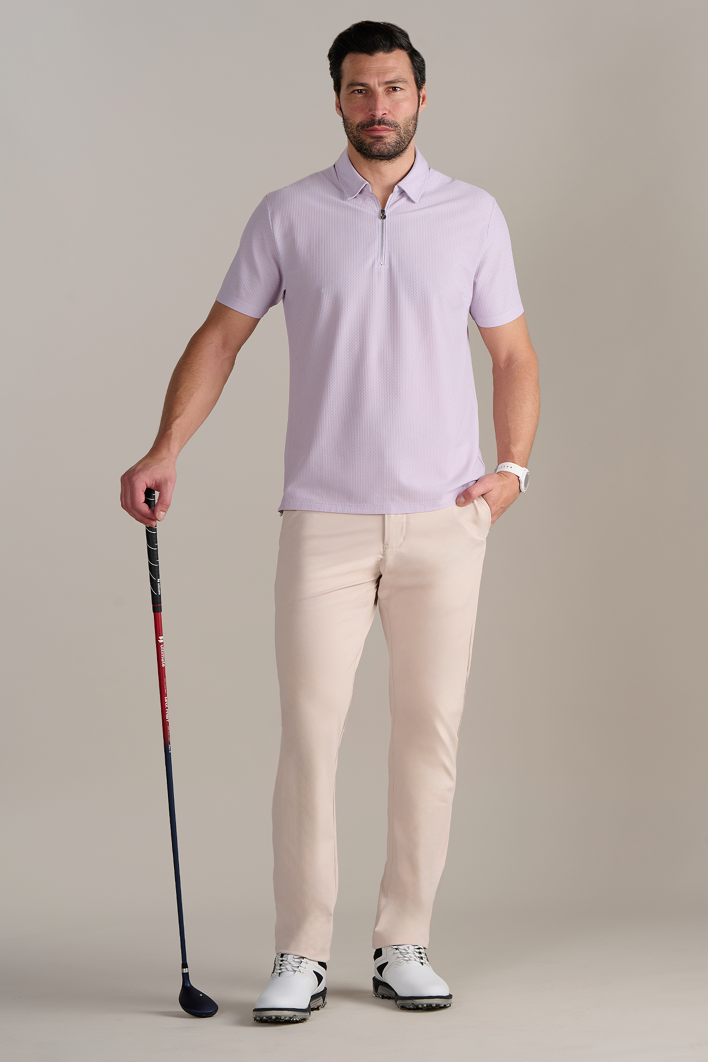A man faces forward against a neutral background, holding a golf club. He wears the Twillory AIR Zip Polo Solid in Lilac, light beige pants, a white watch, and white golf shoes with black accents.