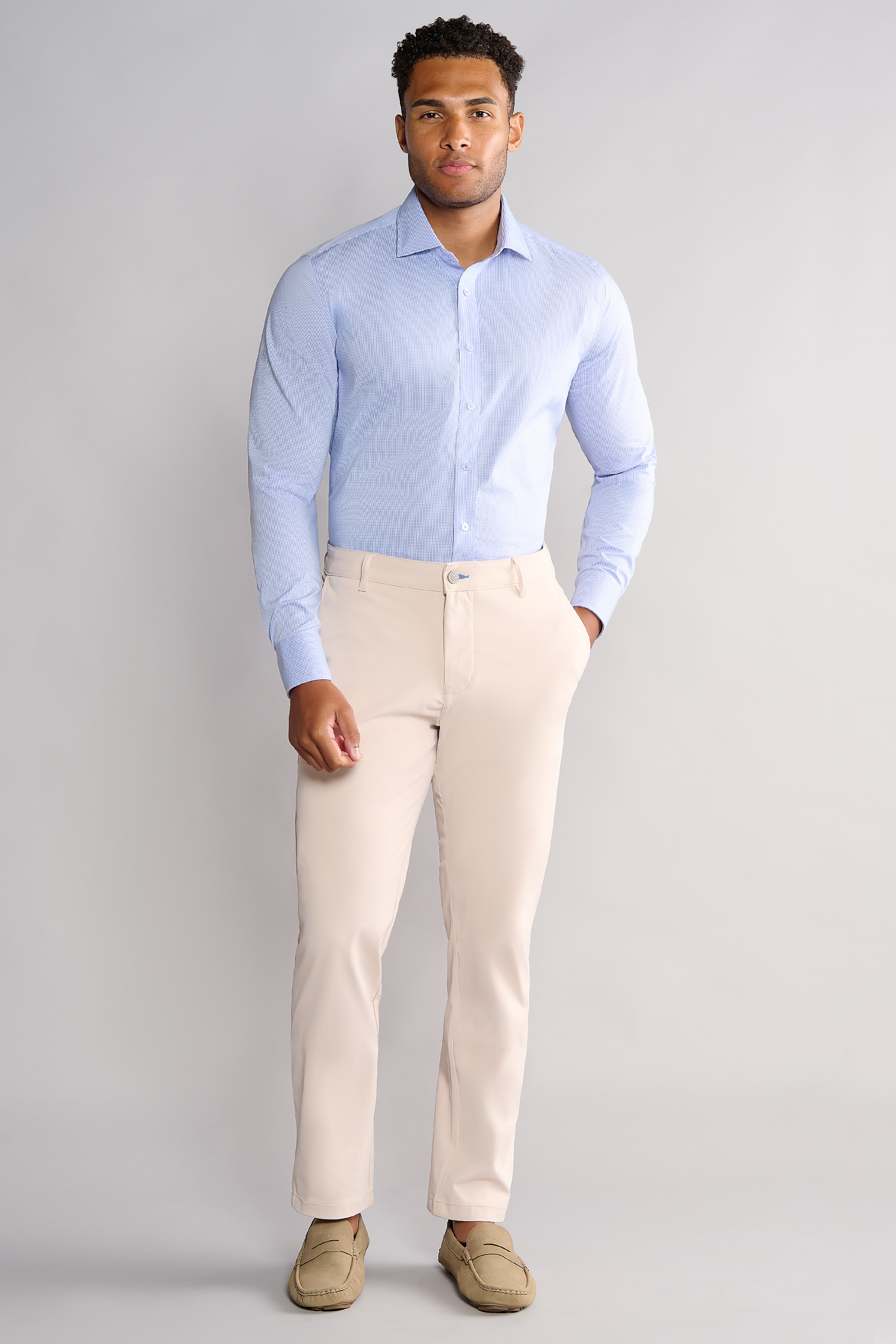 A man stands facing forward with hands in his pockets, wearing the Twillory Leader Houndstooth // Micro Blue shirt, beige dress pants, and tan loafers. His expression is neutral and the background is plain light grey.
