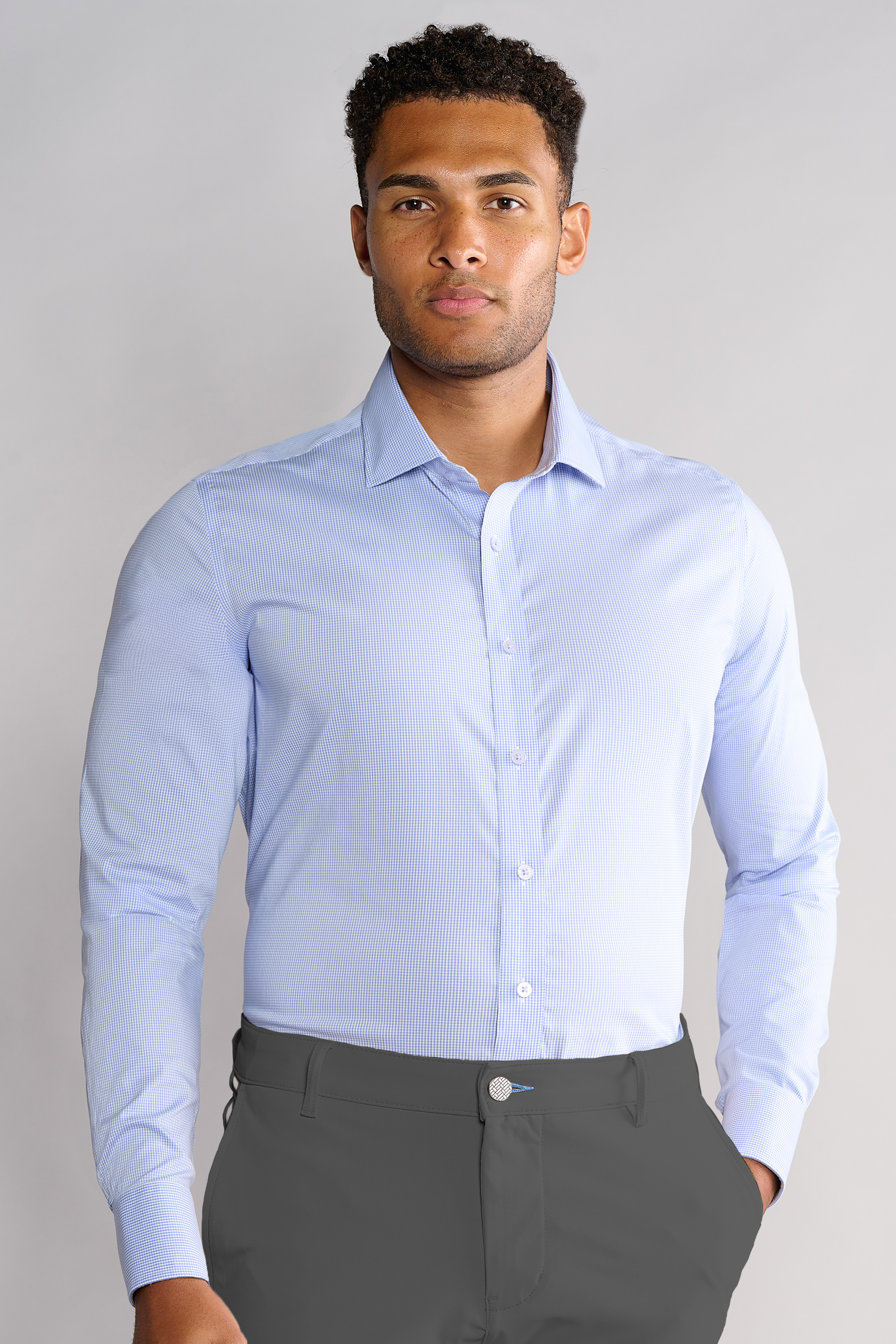 A man with short curly hair stands against a plain light gray background, wearing Twillory's Leader Houndstooth // Micro Blue performance shirt and dark gray pants. The light blue, long-sleeve shirt features 4-way stretch for comfort.
