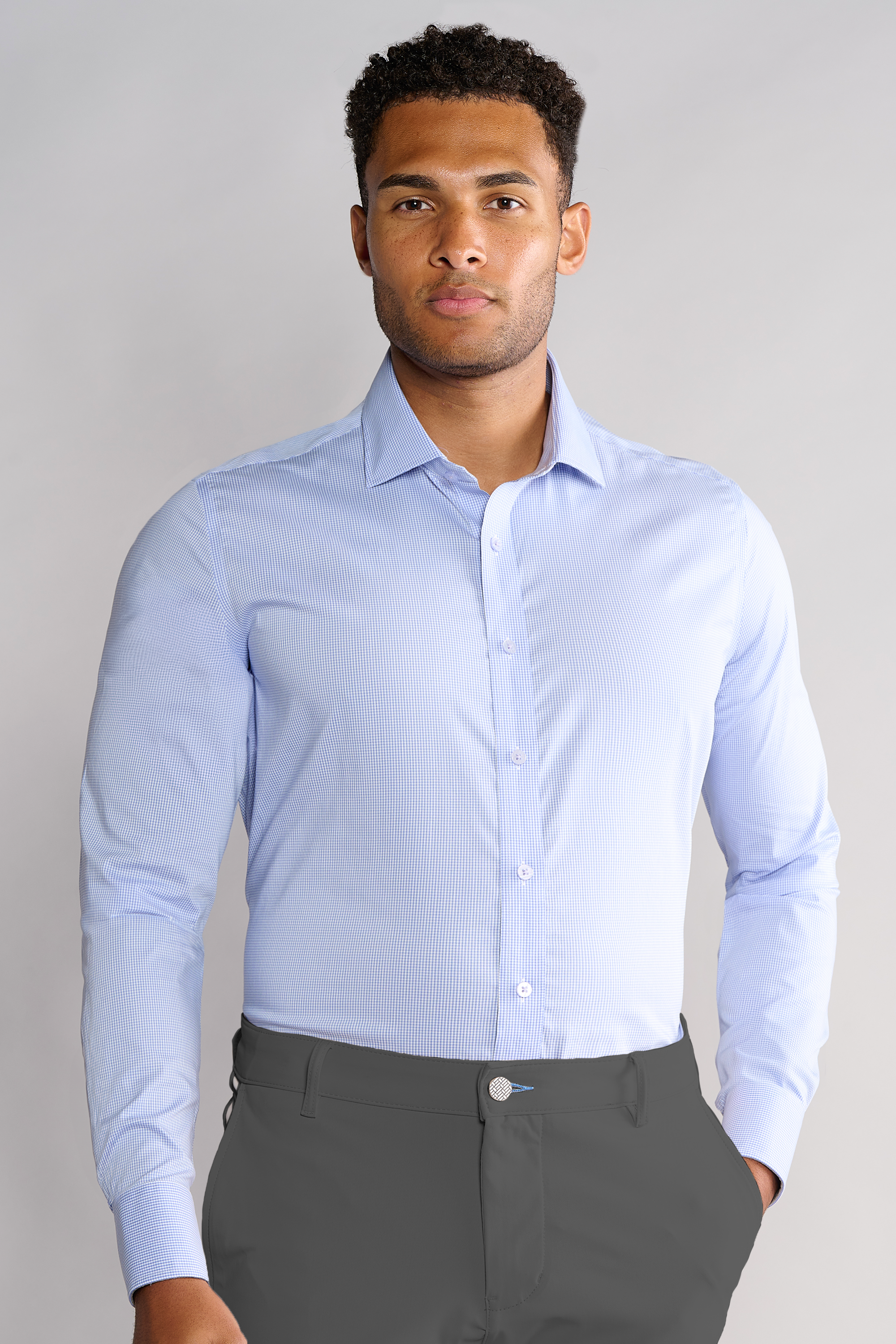 A man with short curly hair stands against a plain light gray background, wearing Twillory's Leader Houndstooth // Micro Blue performance shirt and dark gray pants. The light blue, long-sleeve shirt features 4-way stretch for comfort.