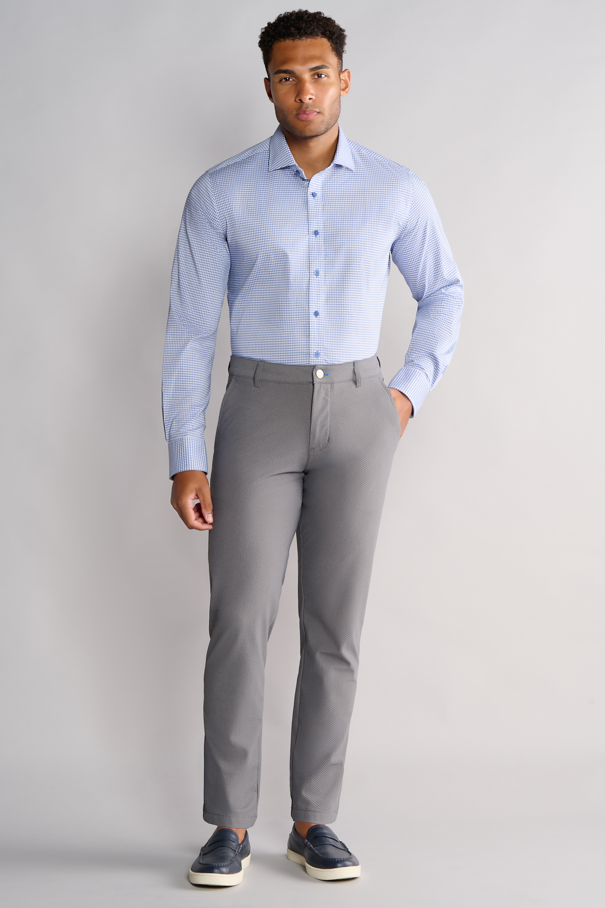 A man stands against a light gray background wearing the Twillory Leader Check // Micro Blue Performance Shirt, featuring 4-way stretch, tucked into slim-fit gray pants. He has dark loafers with white soles and looks at the camera with a neutral expression.