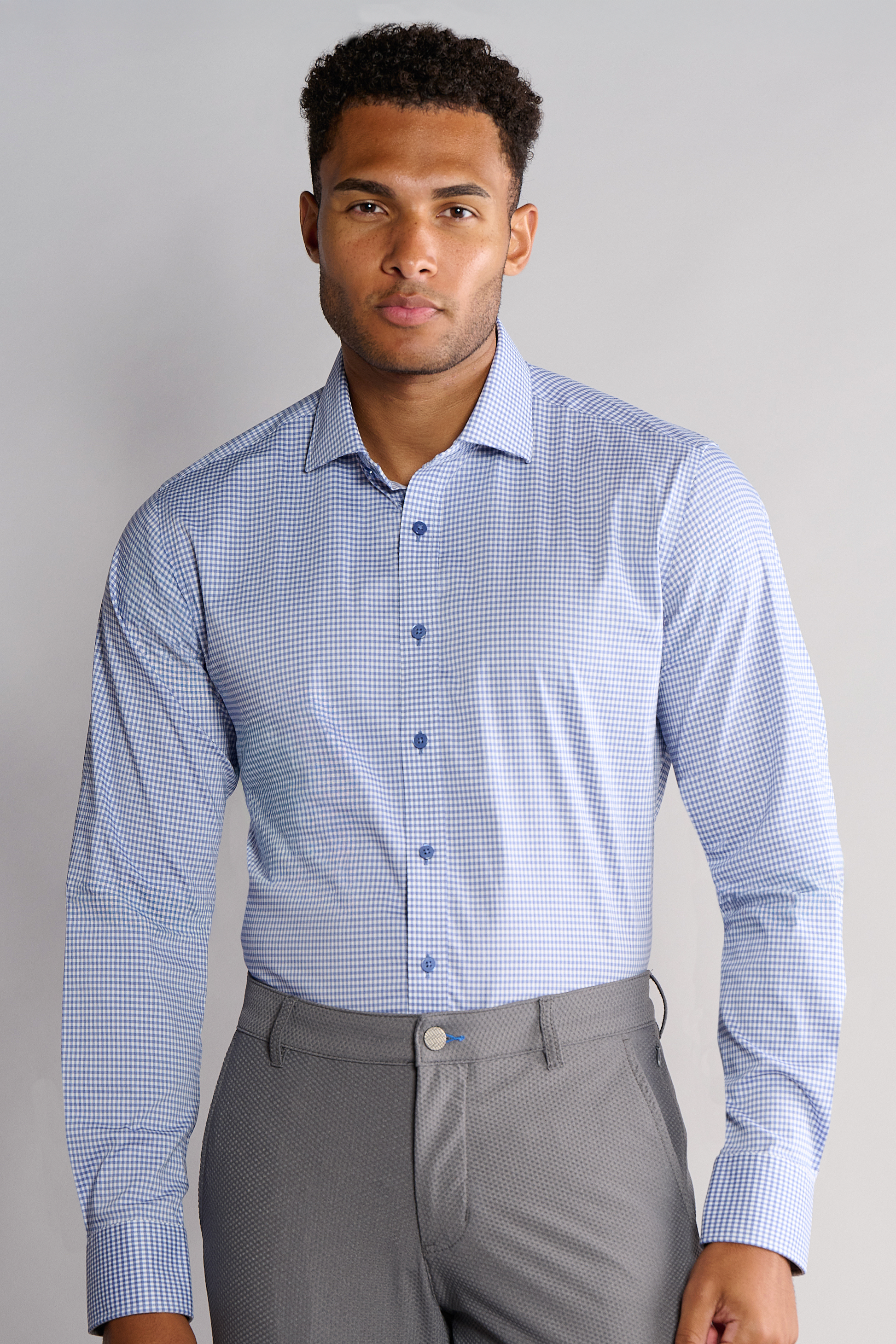 A man faces forward wearing Twillory’s Leader Check // Micro Blue Performance Shirt, featuring small blue checks and blue buttons, neatly tucked into gray dress pants. He appears neutral, set against a plain light gray background.