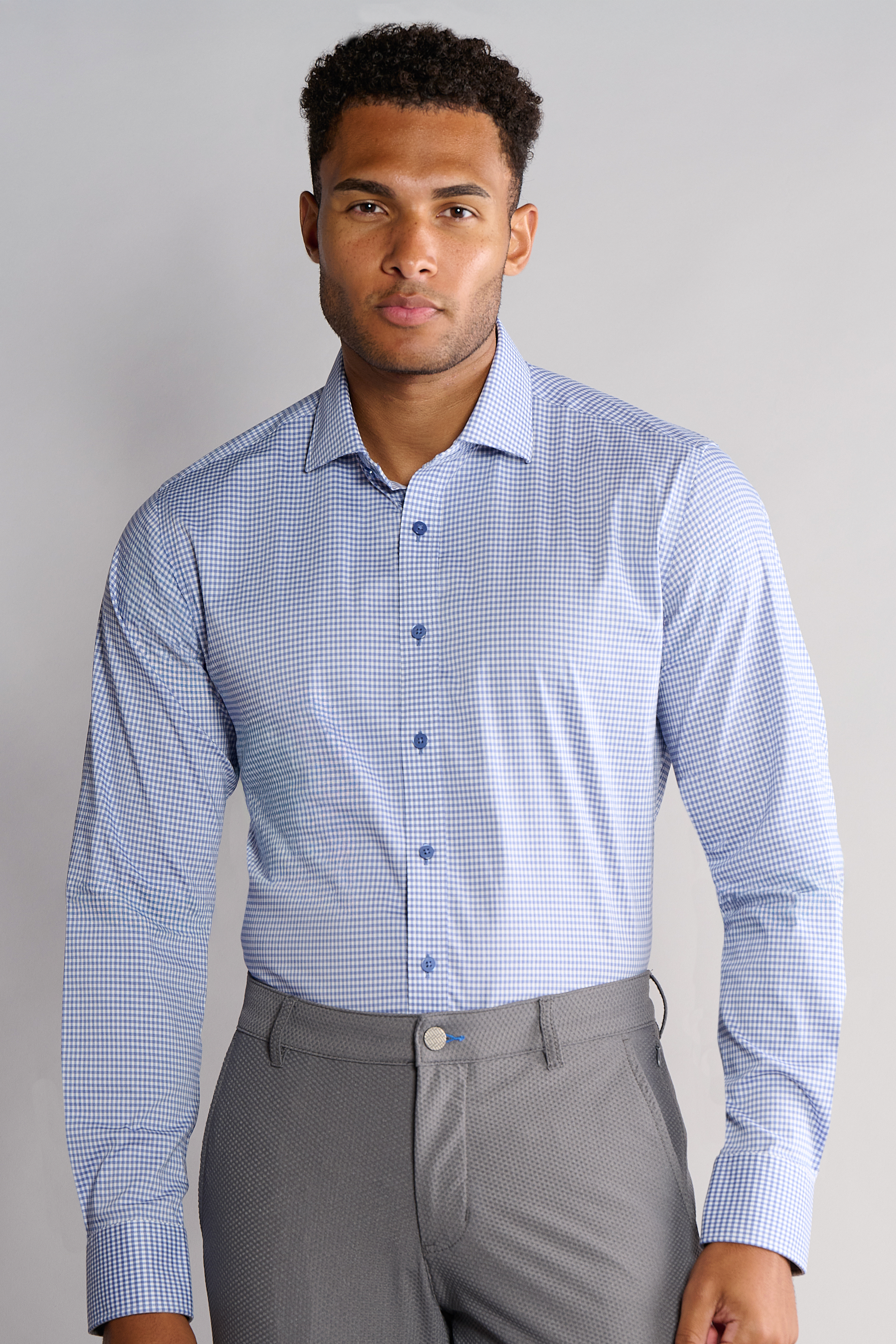 A man faces forward wearing Twillory’s Leader Check // Micro Blue Performance Shirt, featuring small blue checks and blue buttons, neatly tucked into gray dress pants. He appears neutral, set against a plain light gray background.