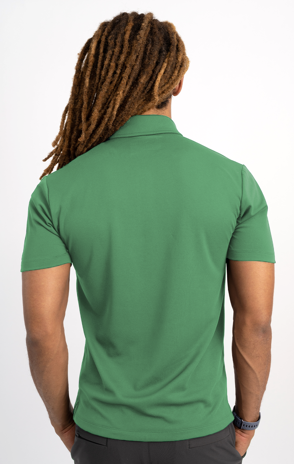 A person with long, thick brown dreadlocks stands facing away from the camera in a mint Short Sleeve Polo Solid by Twillory, paired with dark pants and a black wristwatch, against a plain white background.