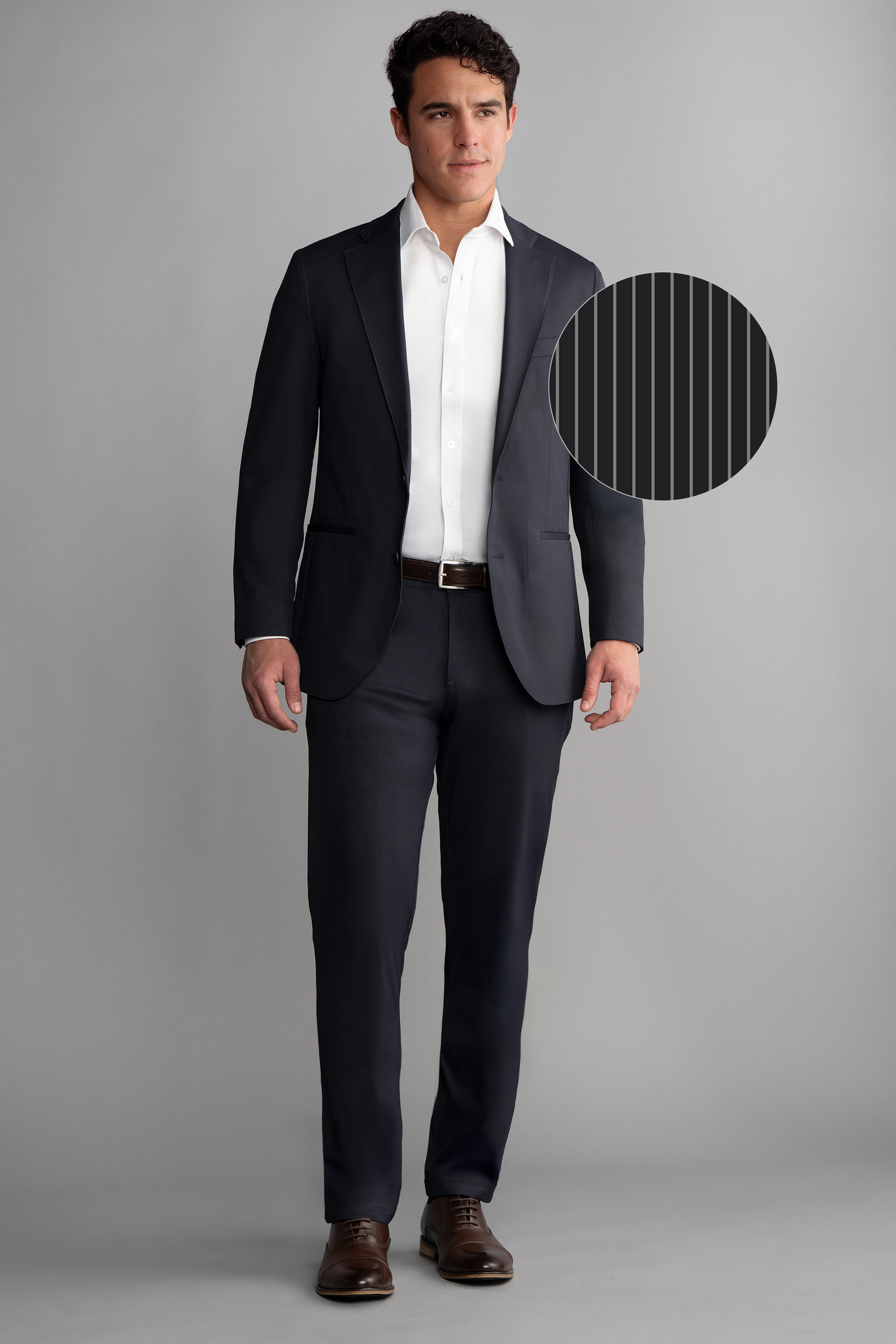 A man stands against a gray backdrop wearing Twillory's Performance Blazer // Navy Pin Stripe, paired with a white dress shirt, brown belt, and brown dress shoes. A circular inset reveals the blazer’s fine vertical pinstripes.
