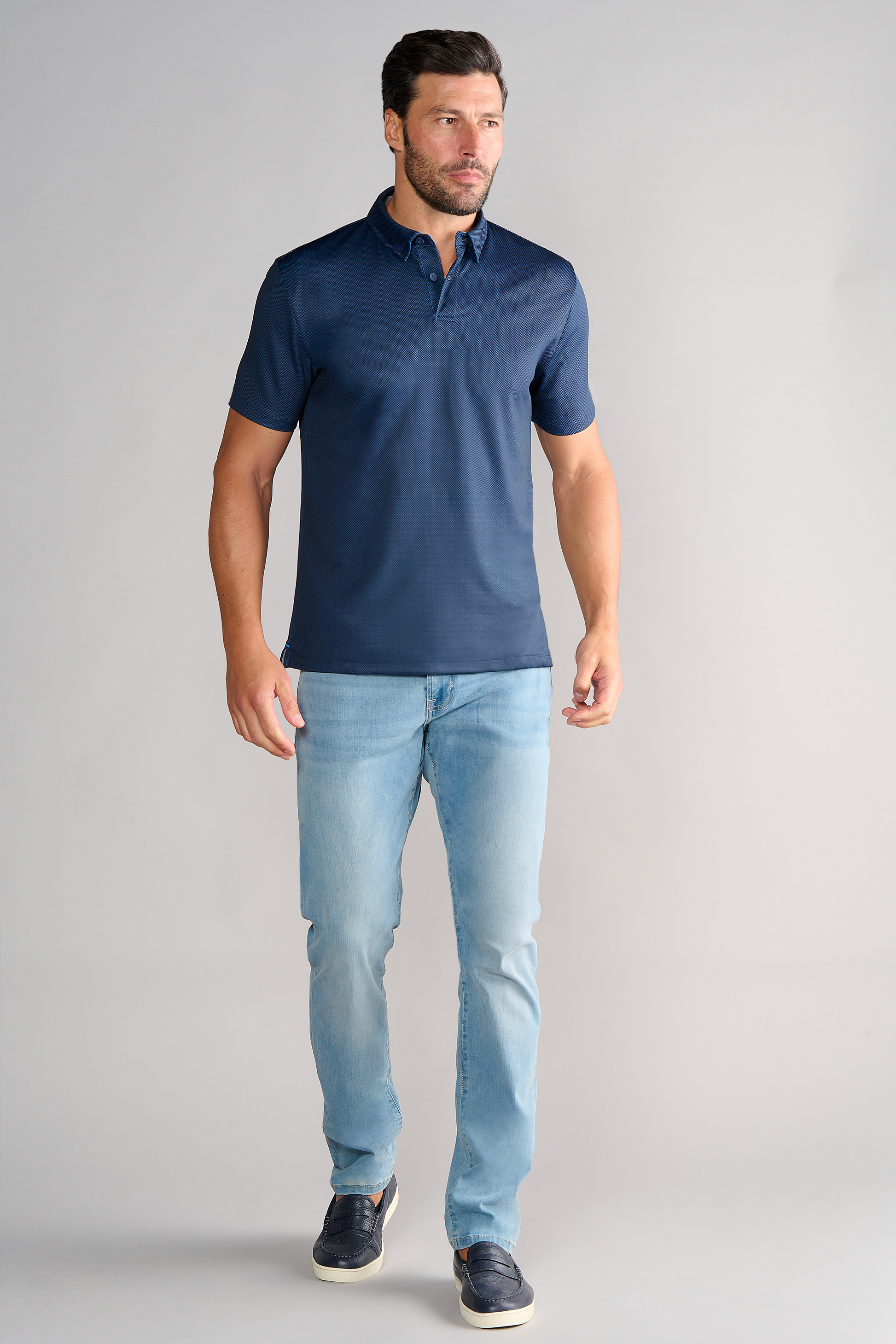 A man with short dark hair and a beard stands against a plain light gray background wearing the Twillory Short Sleeve Polo Dot in Navy, light blue jeans, and dark sneakers. His arms are relaxed at his sides as he looks slightly to his left.