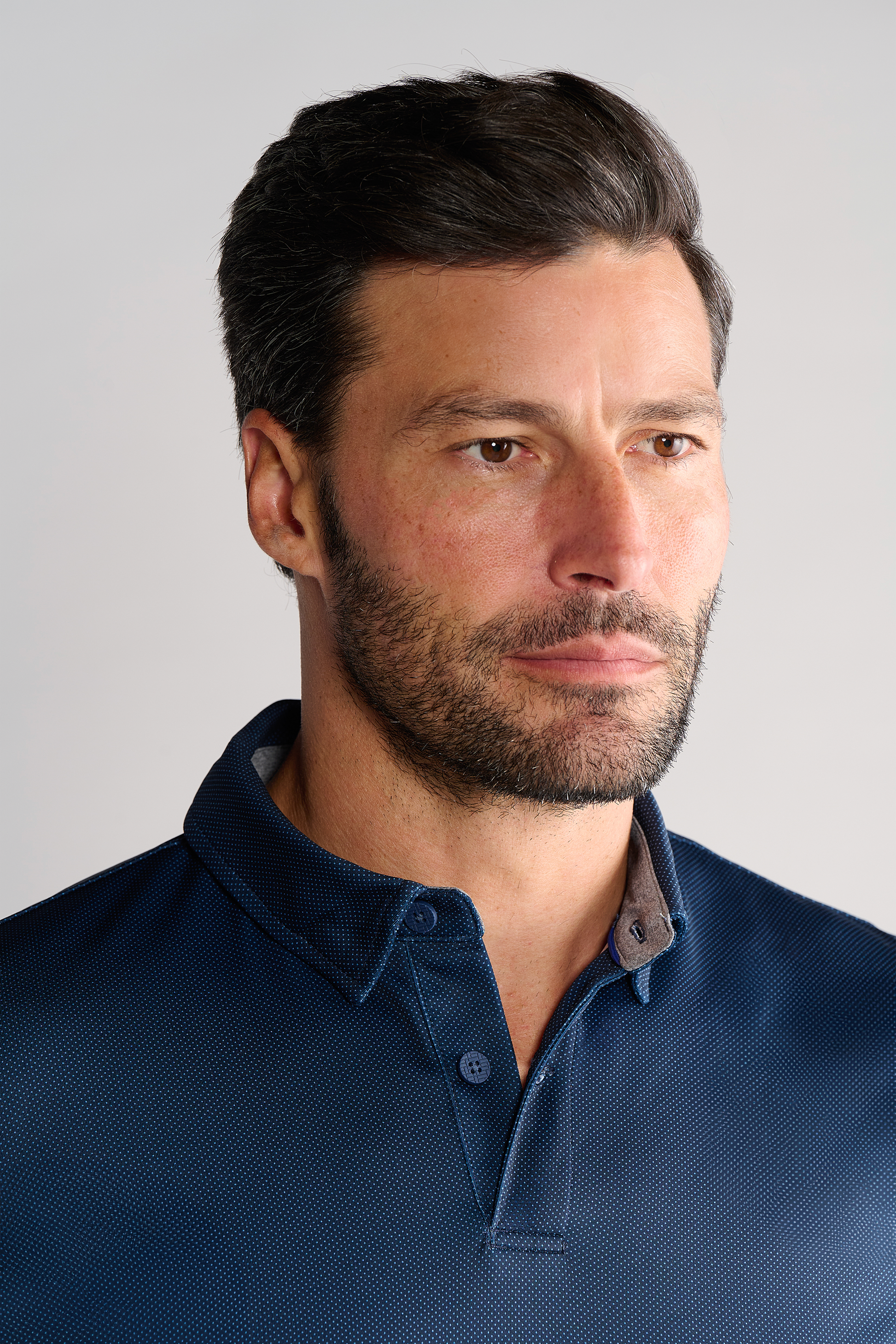 A man with short dark hair and a trimmed beard models the Twillory Short Sleeve Polo Dot in navy. He gazes to the side with a neutral expression, while a plain light backdrop highlights both his features and his stylish shirt.