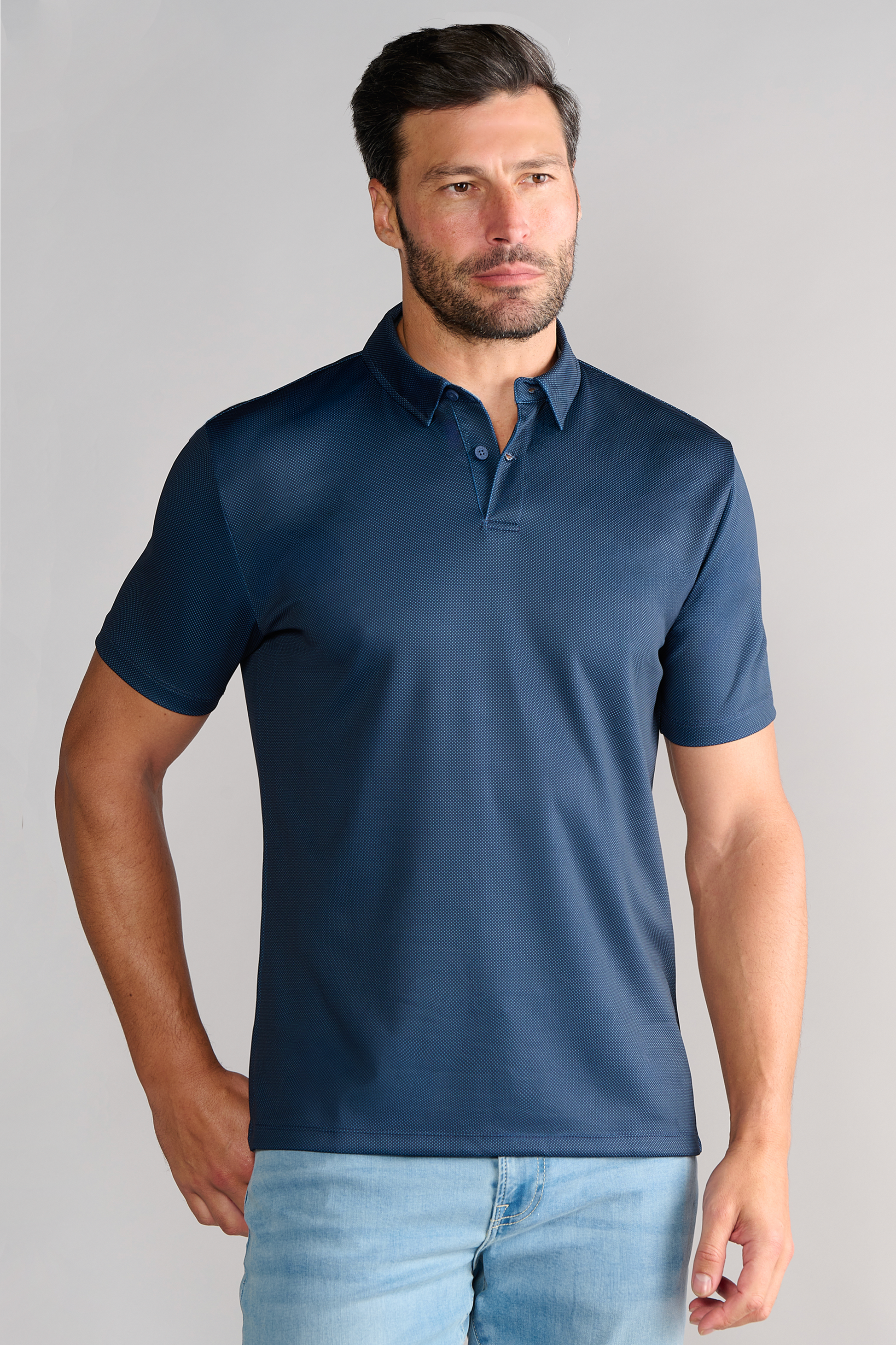 A man with short dark hair and a trimmed beard wears the Twillory Short Sleeve Polo Dot in Navy and light blue jeans, standing against a plain light gray background with a neutral expression, his left hand at his side.
