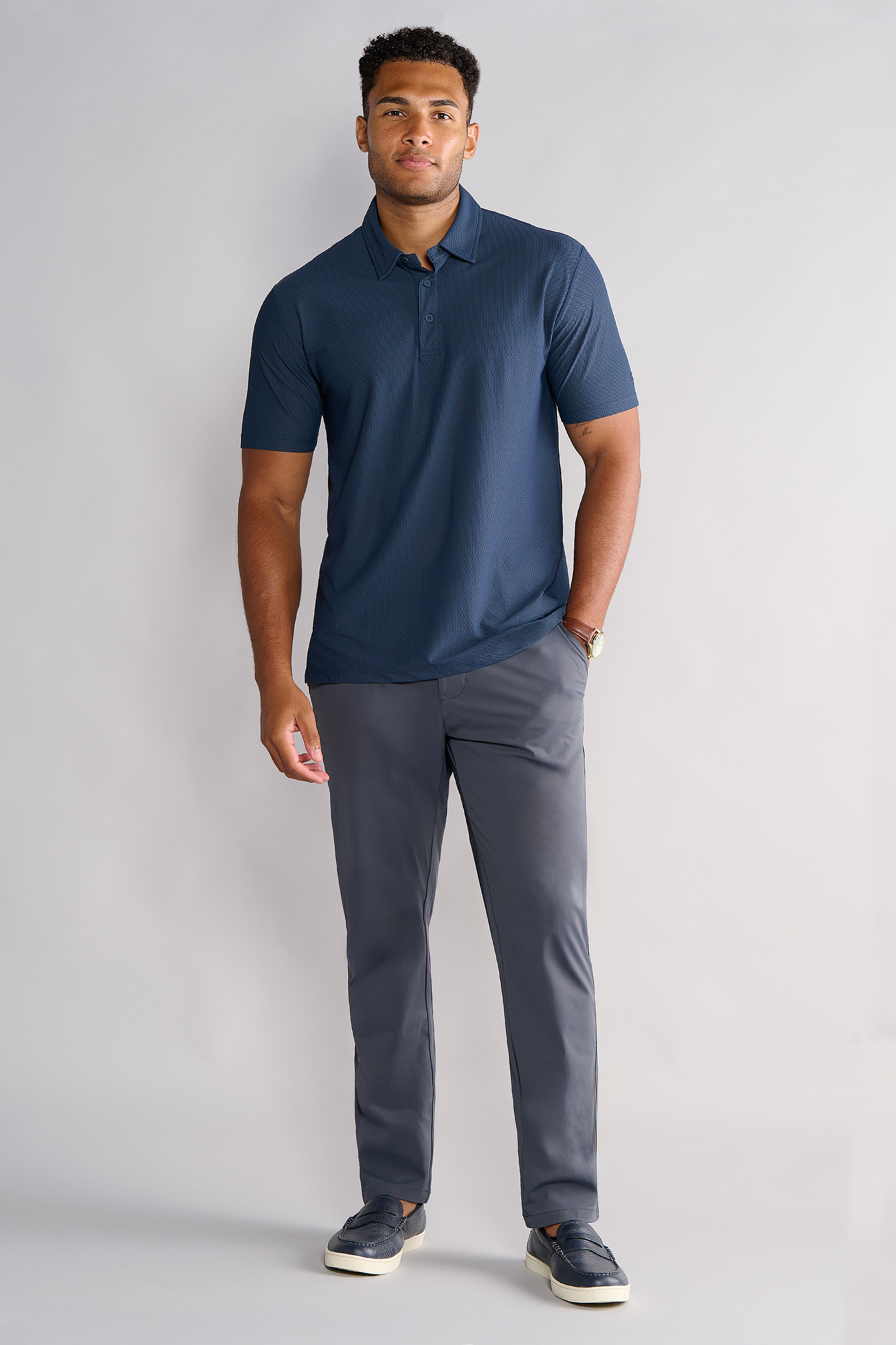 A man with short hair stands against a light gray background, wearing the Twillory AIR 3 Button Polo Solid in Navy, gray dress pants, navy slip-on shoes, and a watch. He appears relaxed and faces forward with a neutral expression.