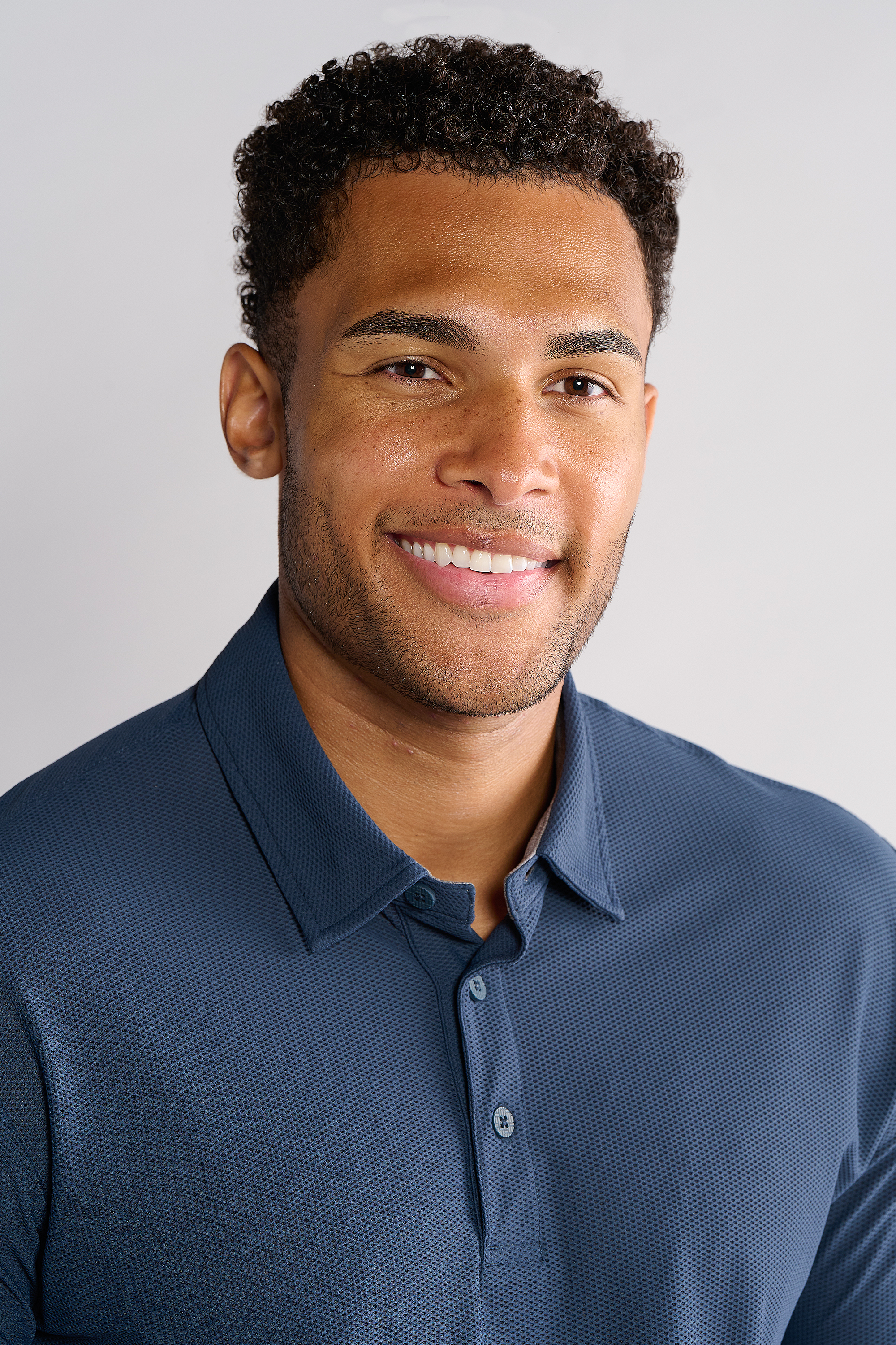 A man with short, curly hair and trimmed facial hair smiles at the camera, wearing Twillory's AIR 3 Button Polo Solid in Navy. The hyper-breathable polo stands out against a plain, light background for a clean, professional look.