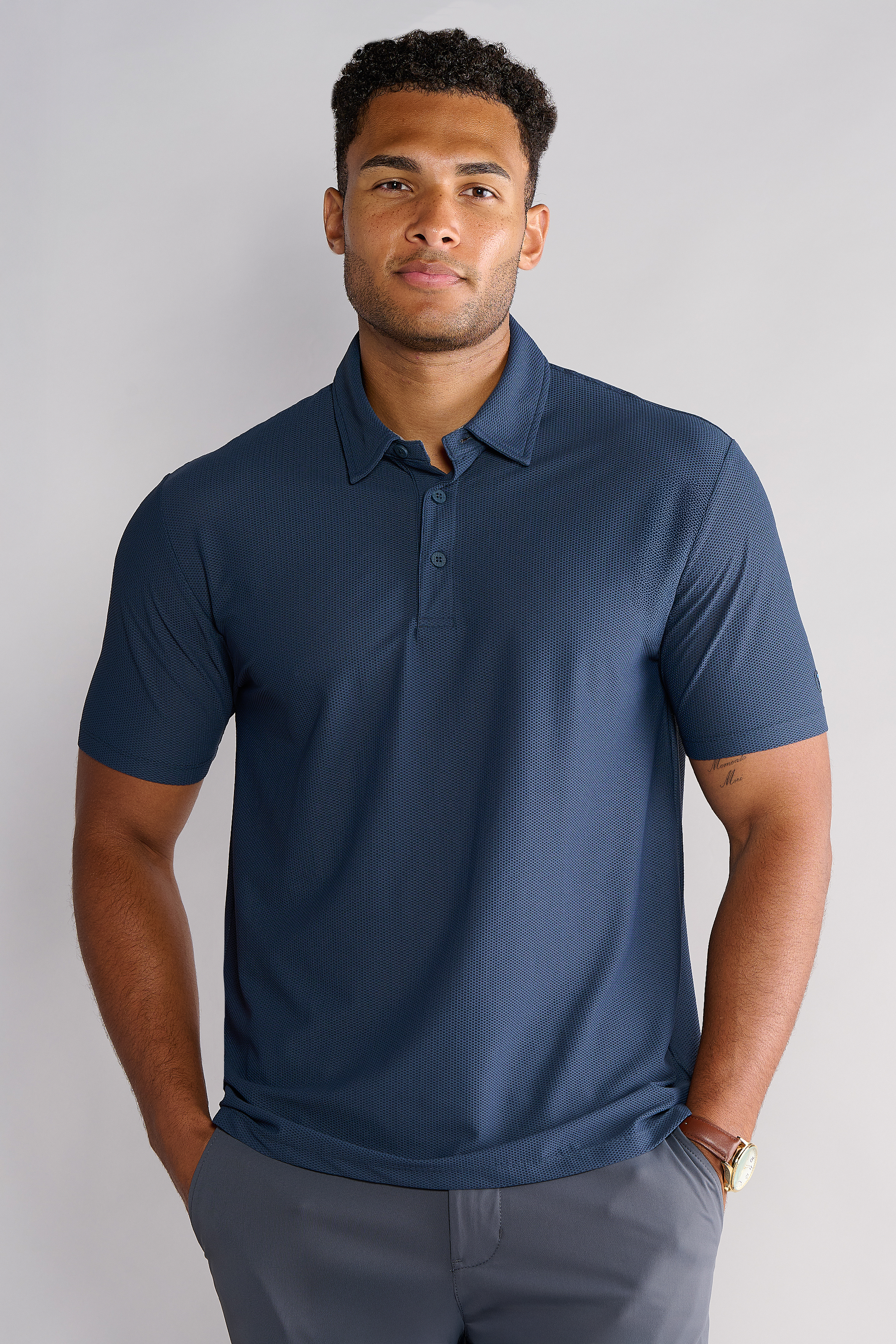 A man with short, curly hair and a neat beard stands against a light gray background, smiling softly at the camera. He's wearing the Twillory AIR 3 Button Polo Solid in Navy and gray pants, with his hands in his pockets and a watch on his left wrist.