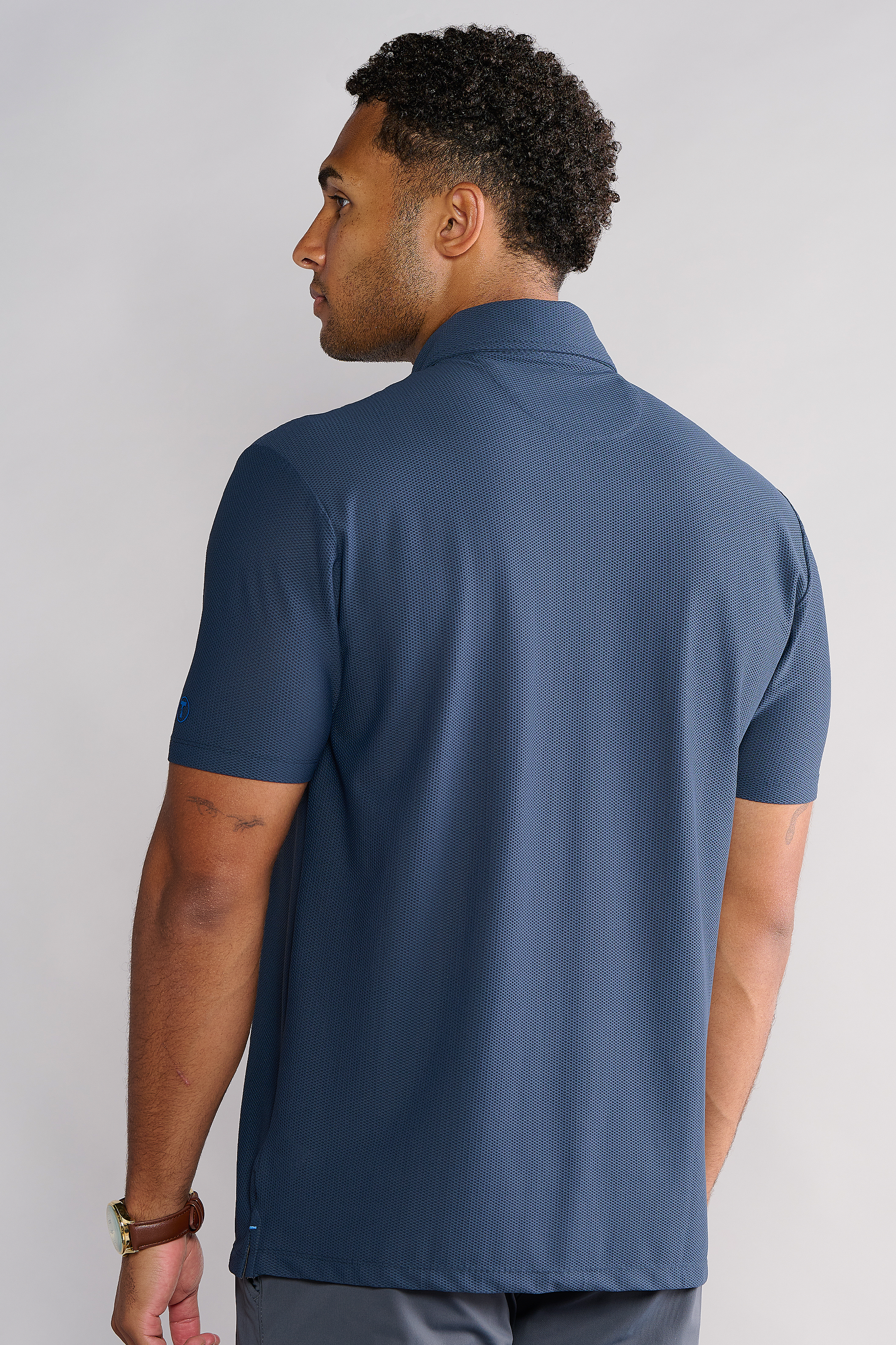 A man with short curly hair, seen from behind, wears the Twillory AIR 3 Button Polo Solid in navy and a brown wristwatch. The polo is crafted from 360° Airflow Fabric for hyper-breathability. The background is light gray.