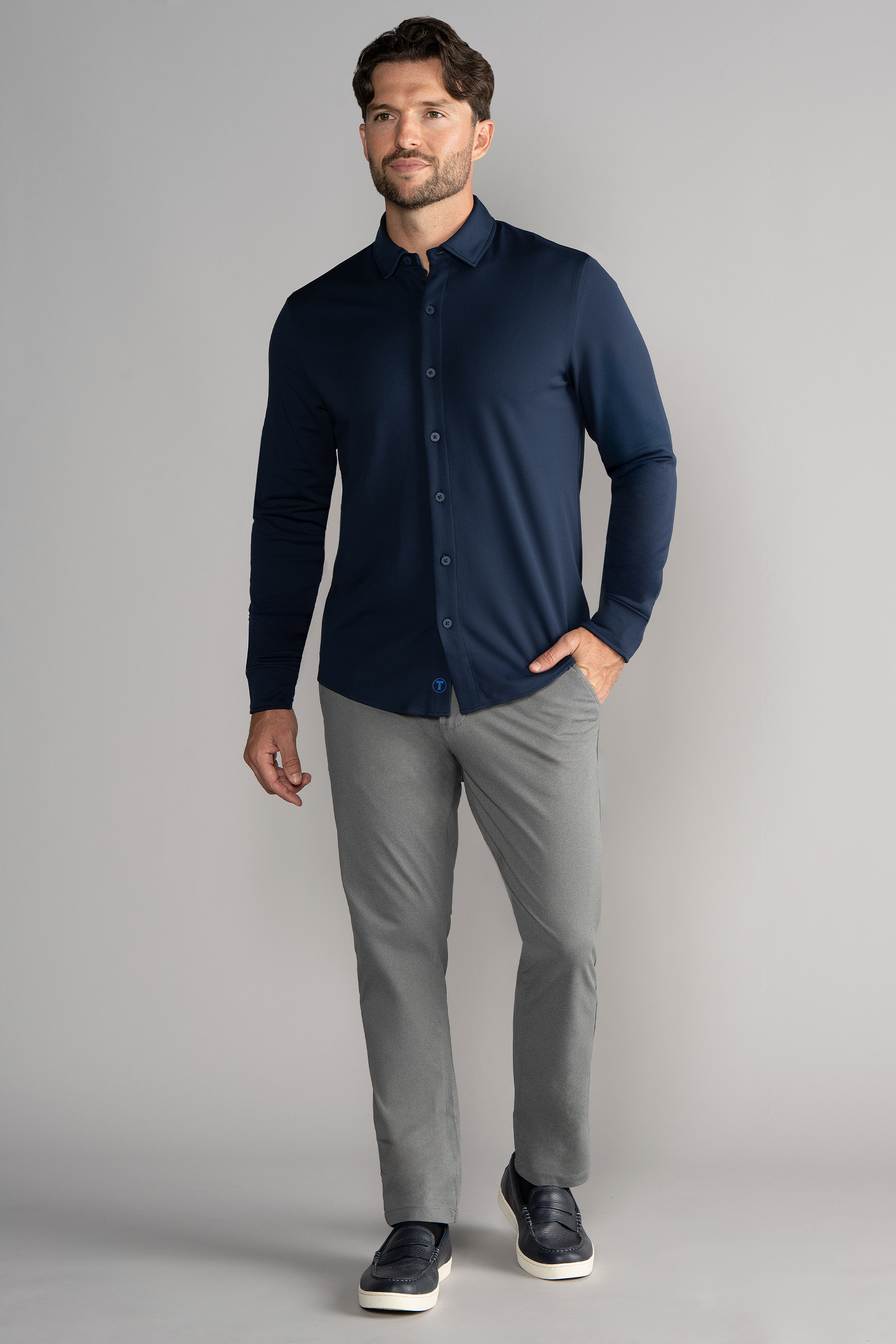 A man with short brown hair and a beard wears the Twillory Long Sleeve Button Down Polo Solid in navy, light gray pants, and dark slip-on shoes. He stands facing forward against a plain light gray background, smiling slightly.