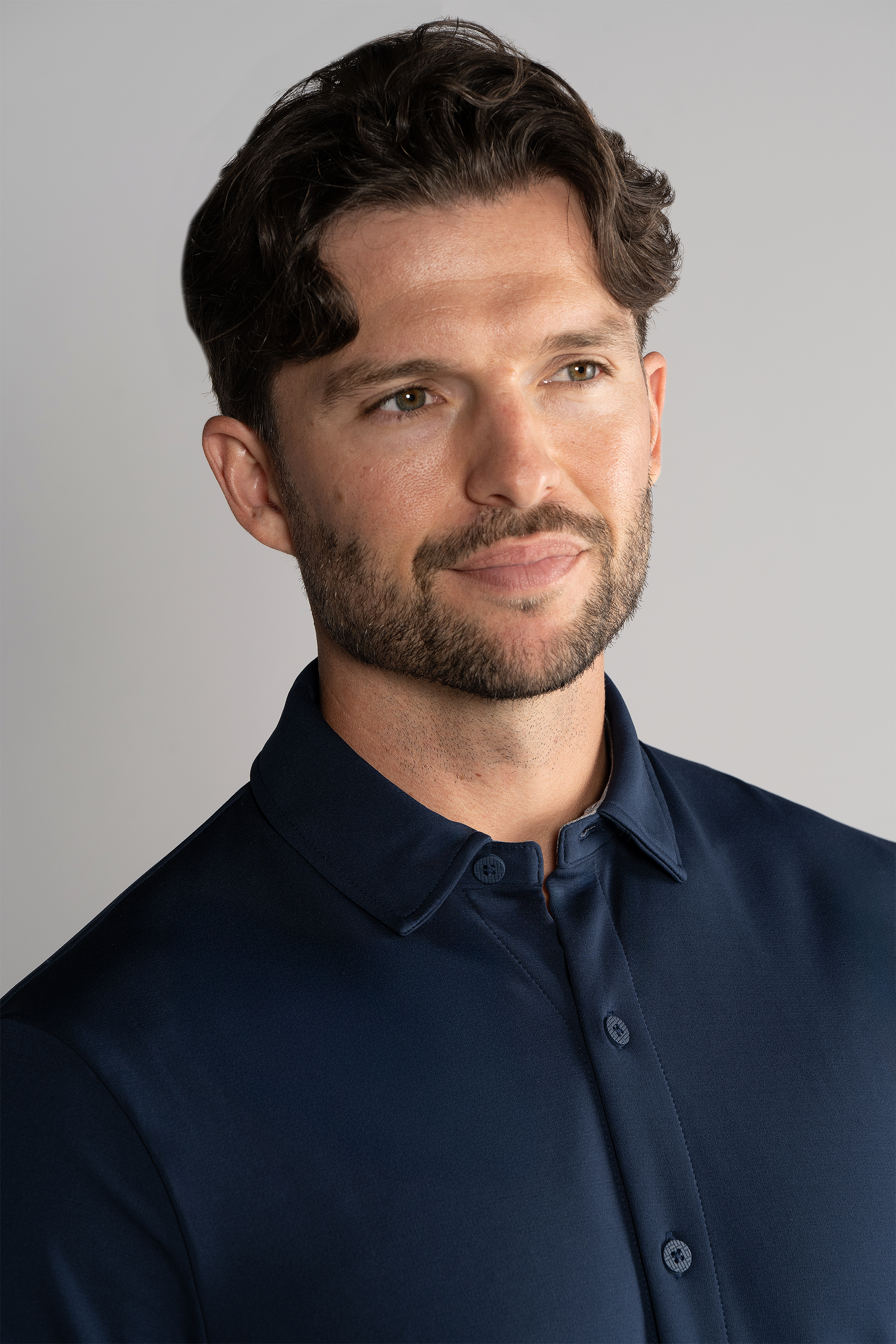 A man with short, wavy brown hair and a trimmed beard smiles gently while wearing the Twillory Long Sleeve Button Down Polo Solid in Navy, set against a plain light gray background.