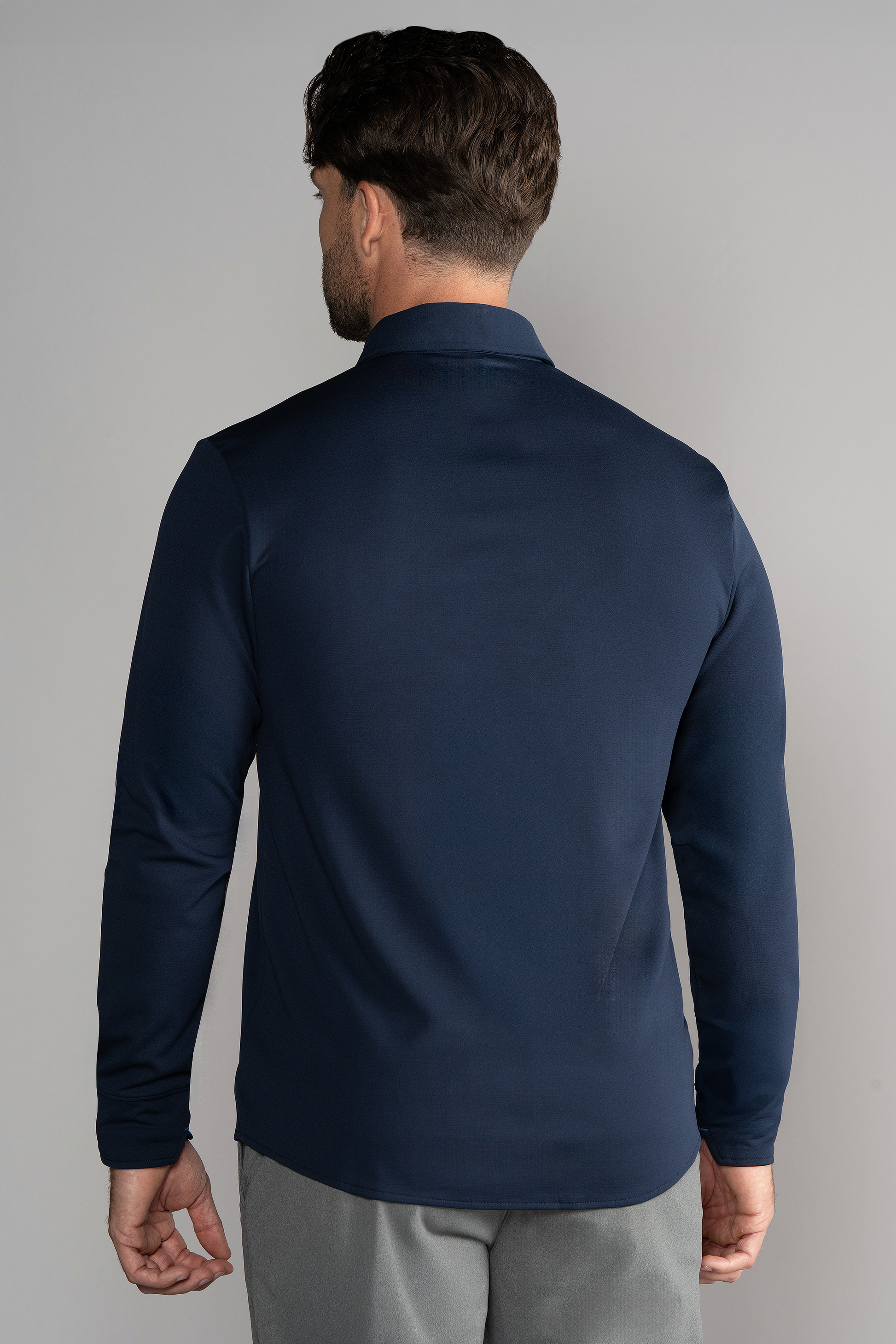 A man stands facing away from the camera against a plain gray background, wearing Twillory's Long Sleeve Button Down Polo Solid in Navy and light gray pants. He has short dark brown hair, with his arms resting naturally by his sides.