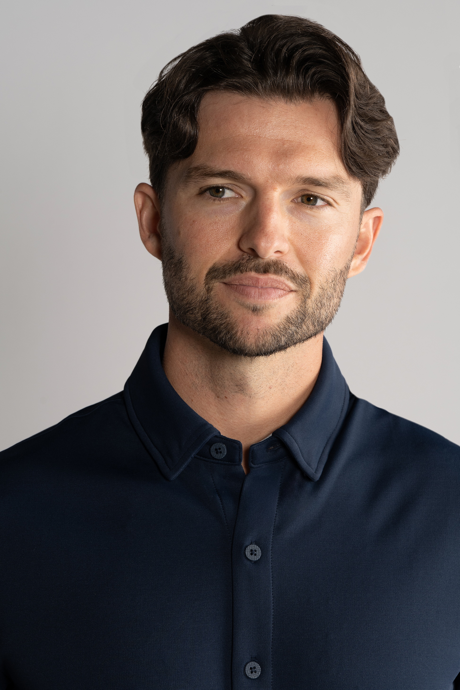 A man with short brown wavy hair and a trimmed beard wears the Twillory Short Sleeve Button Down Polo Solid in Navy. He smiles softly, looking slightly left, against a plain light gray background showing his head and upper torso.