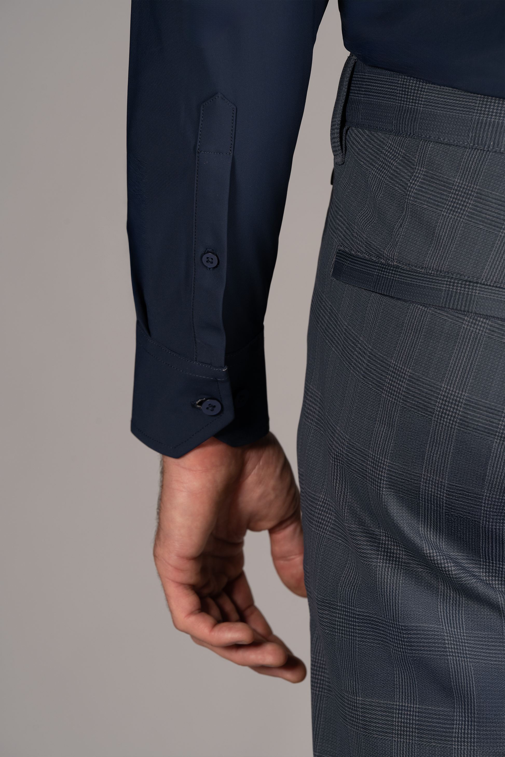A close-up of a person’s left arm and hip showcases Twillory’s The Event Shirt // Navy, a wrinkle-free officewear piece with buttoned cuffs, paired with gray plaid dress pants against a plain light gray background.