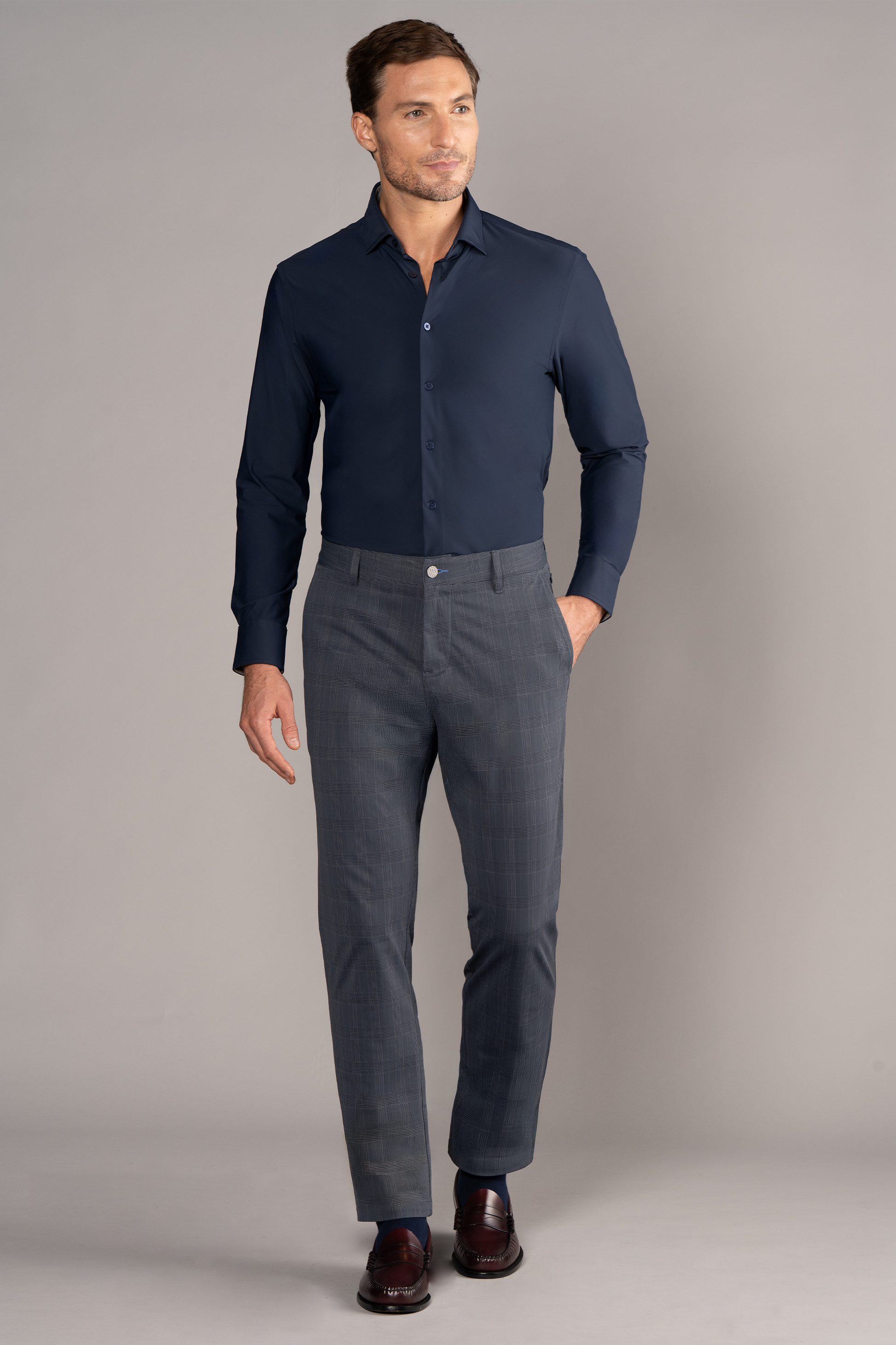 A man with short hair and a trimmed beard stands against a gray background, smiling slightly to his left while wearing Twillory’s The Event Shirt in Navy, paired with gray plaid dress pants and dark brown dress shoes.