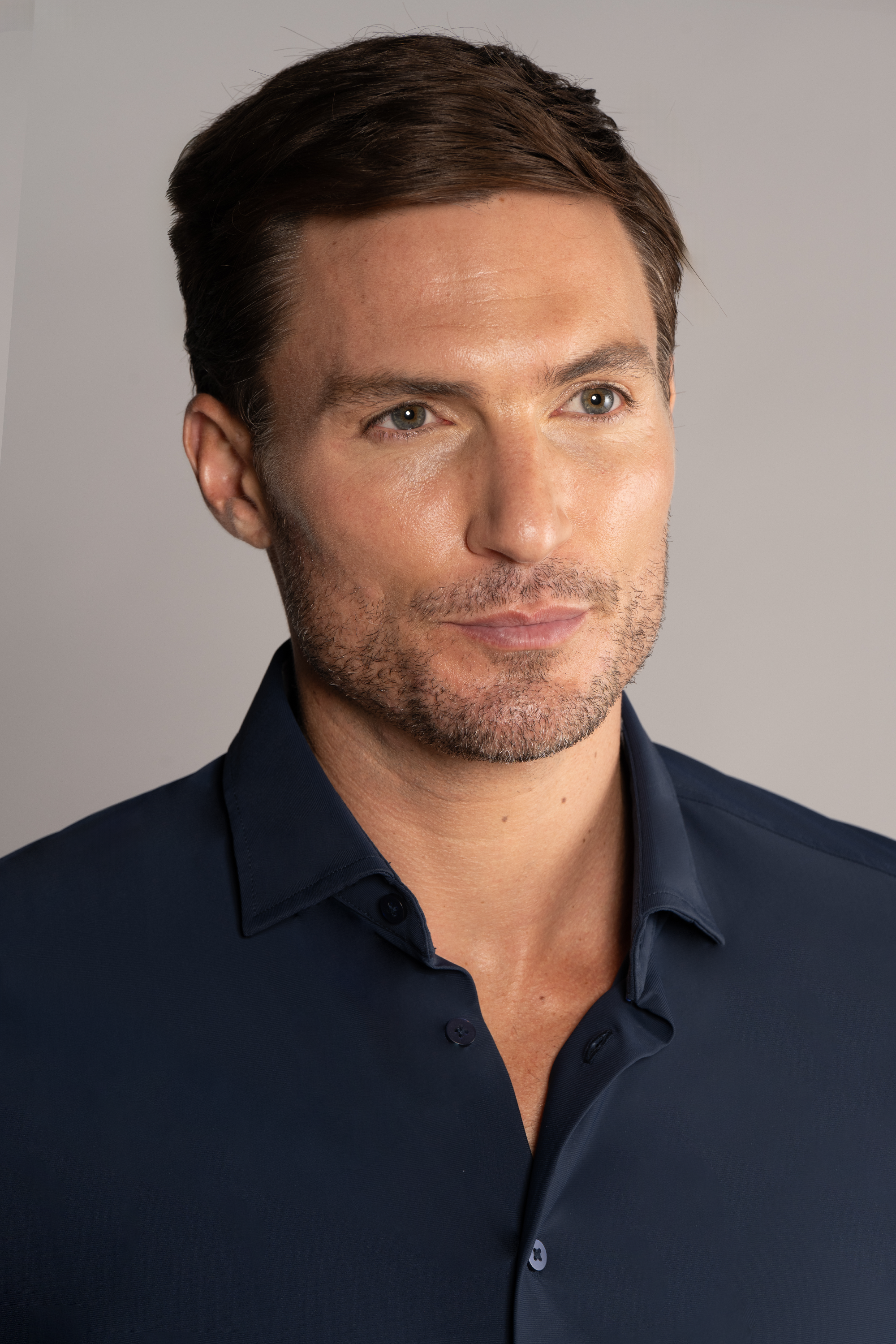 A man with short brown hair and light stubble wears Twillory’s The Event Shirt // Navy, a wrinkle-free piece ideal for the office. Facing slightly right with a neutral look, he’s shown against a softly lit plain background highlighting his face and upper torso.