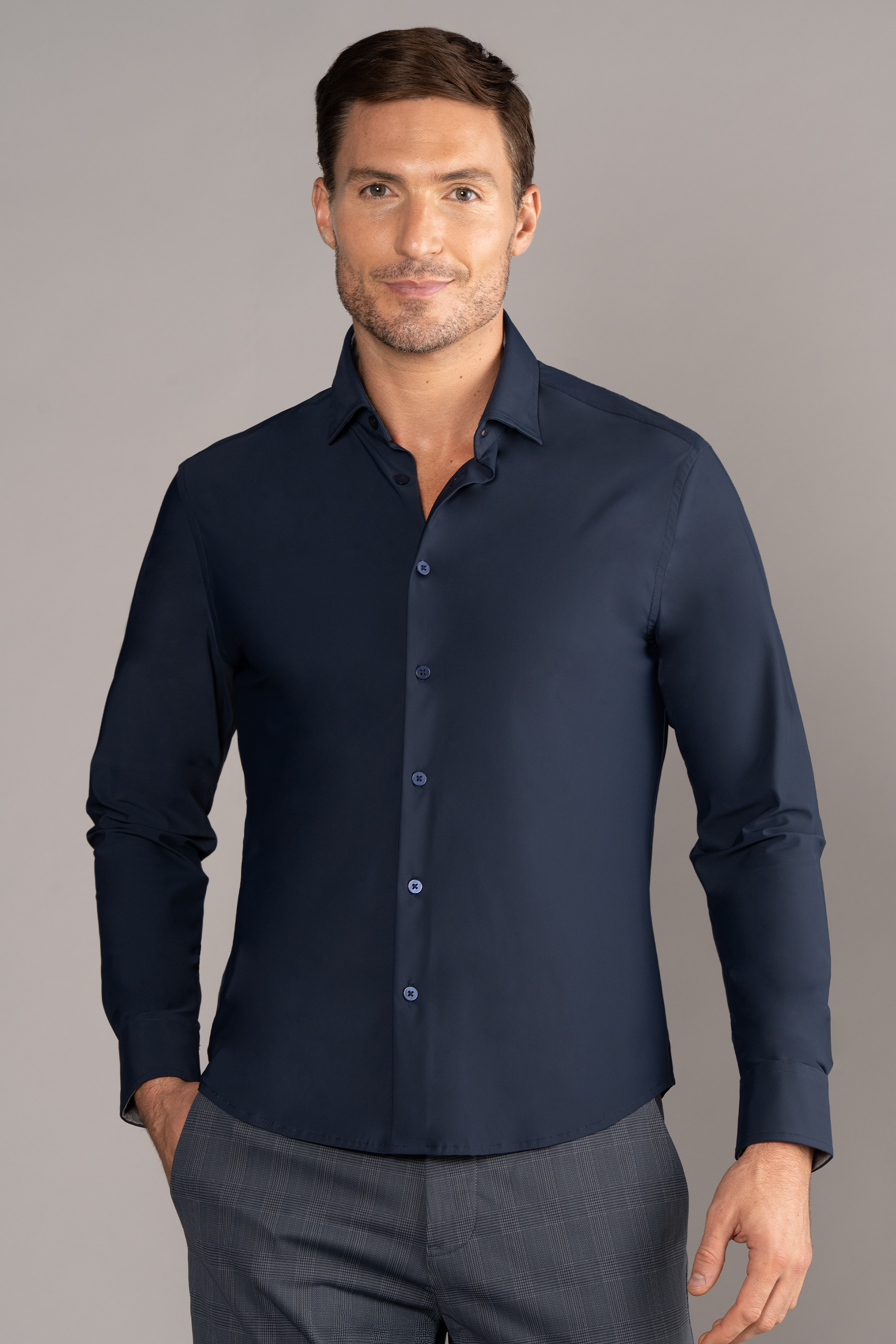 A man with short brown hair and light stubble stands against a gray background, wearing Twillory's The Event Shirt // Navy—fitted, wrinkle-free officewear. He smiles softly at the camera with one hand in his pocket, paired with gray plaid trousers.