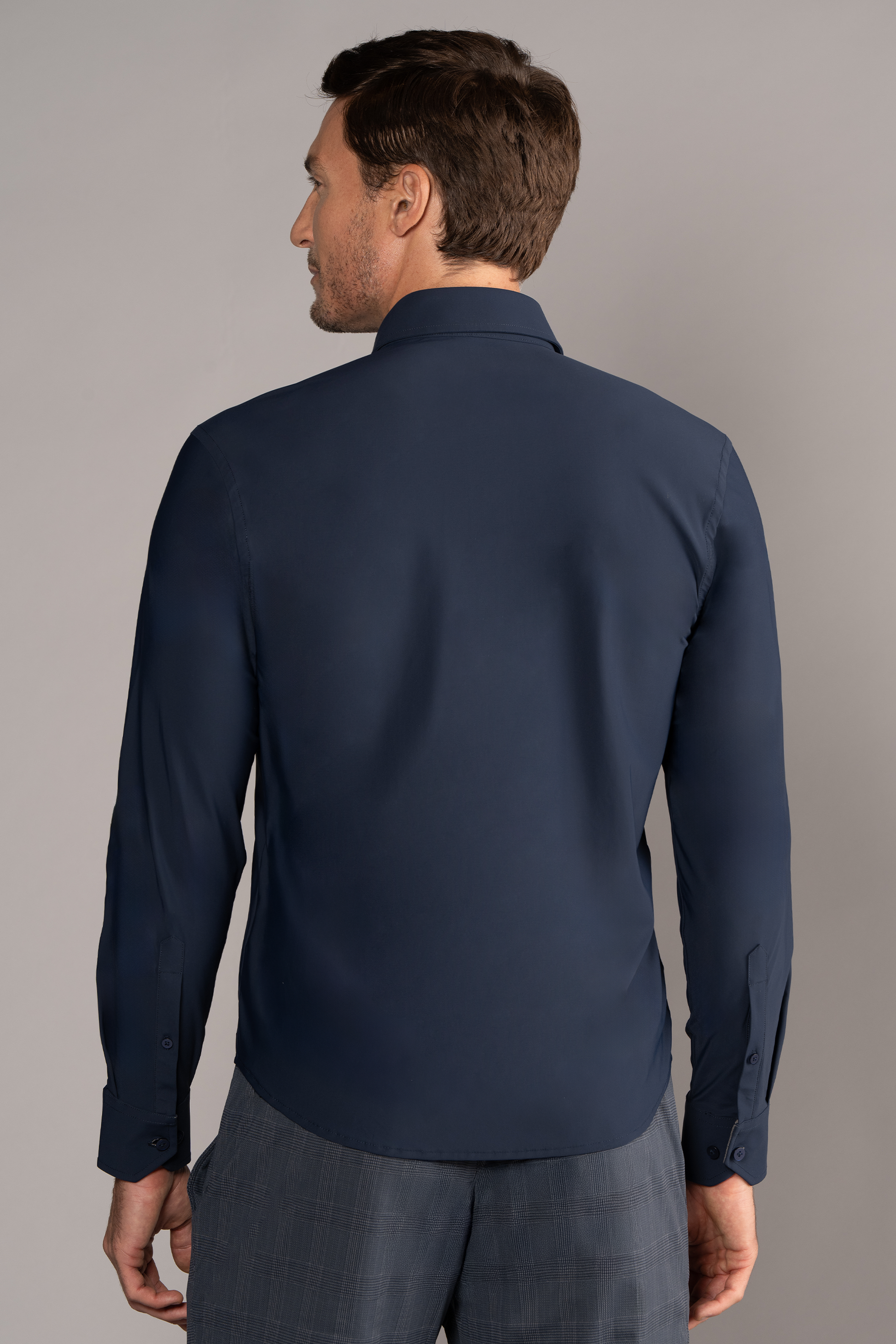 A man with short brown hair, seen from the back, wears Twillory’s The Event Shirt // Navy and gray plaid pants—perfect for modern officewear. He stands against a plain light gray background with his left hand relaxed by his side.