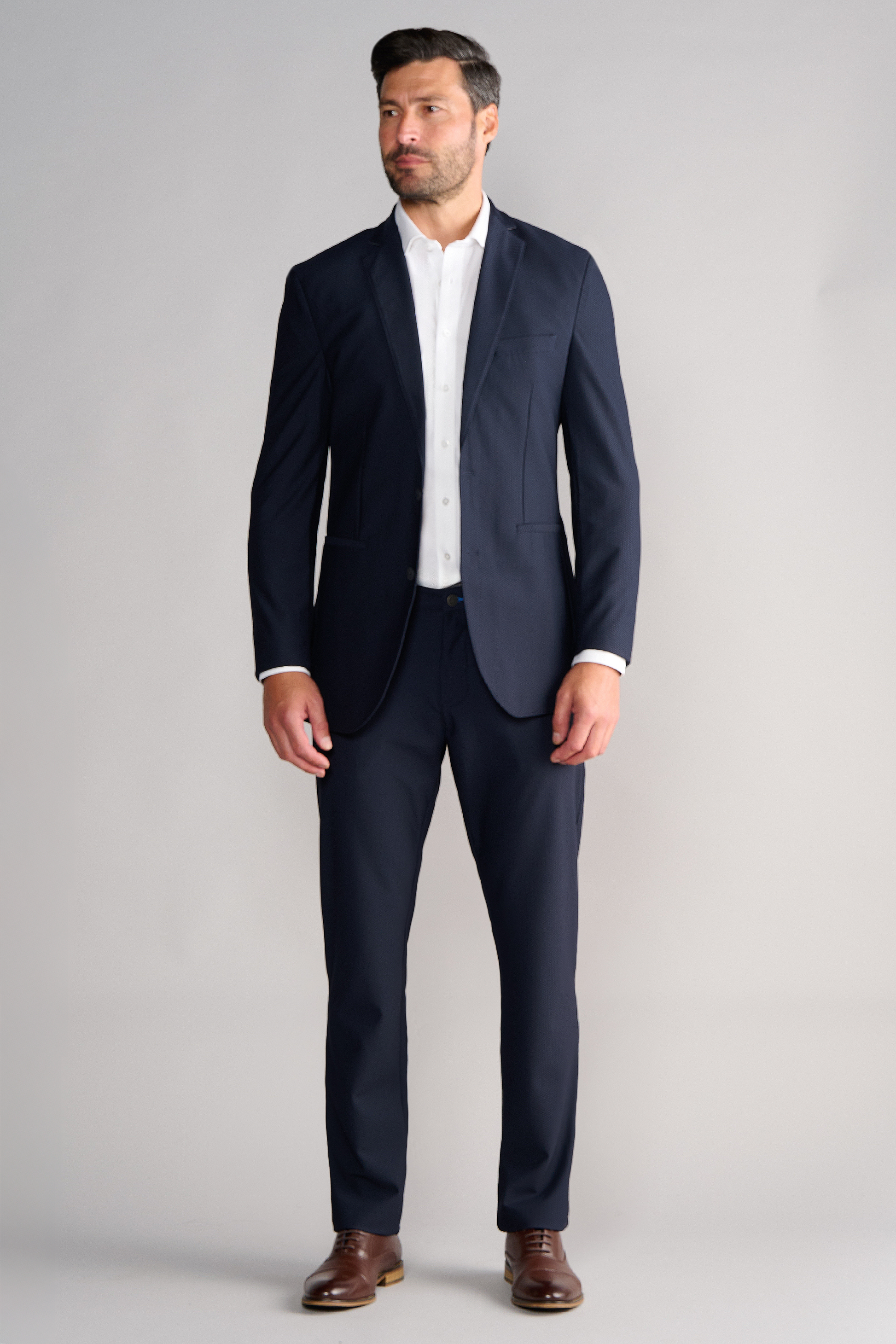 A man faces forward in a Twillory AIR Blazer // Navy over a white dress shirt, no tie, short dark hair and beard, brown dress shoes, all set against a plain light gray backdrop.
