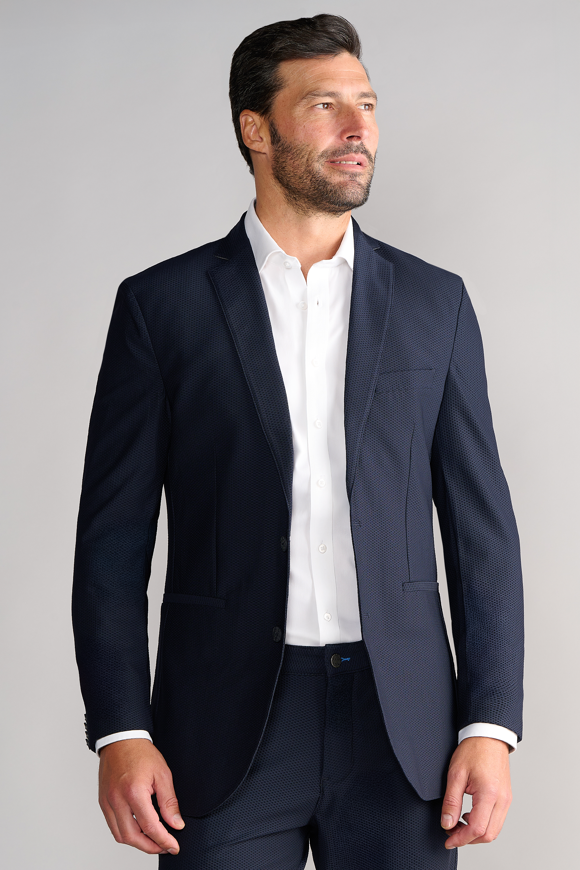 A man with short dark hair and a trimmed beard wears Twillory's AIR Blazer in Navy over a white dress shirt. He faces slightly left with his right hand at his side, looking thoughtful against a plain light gray background.
