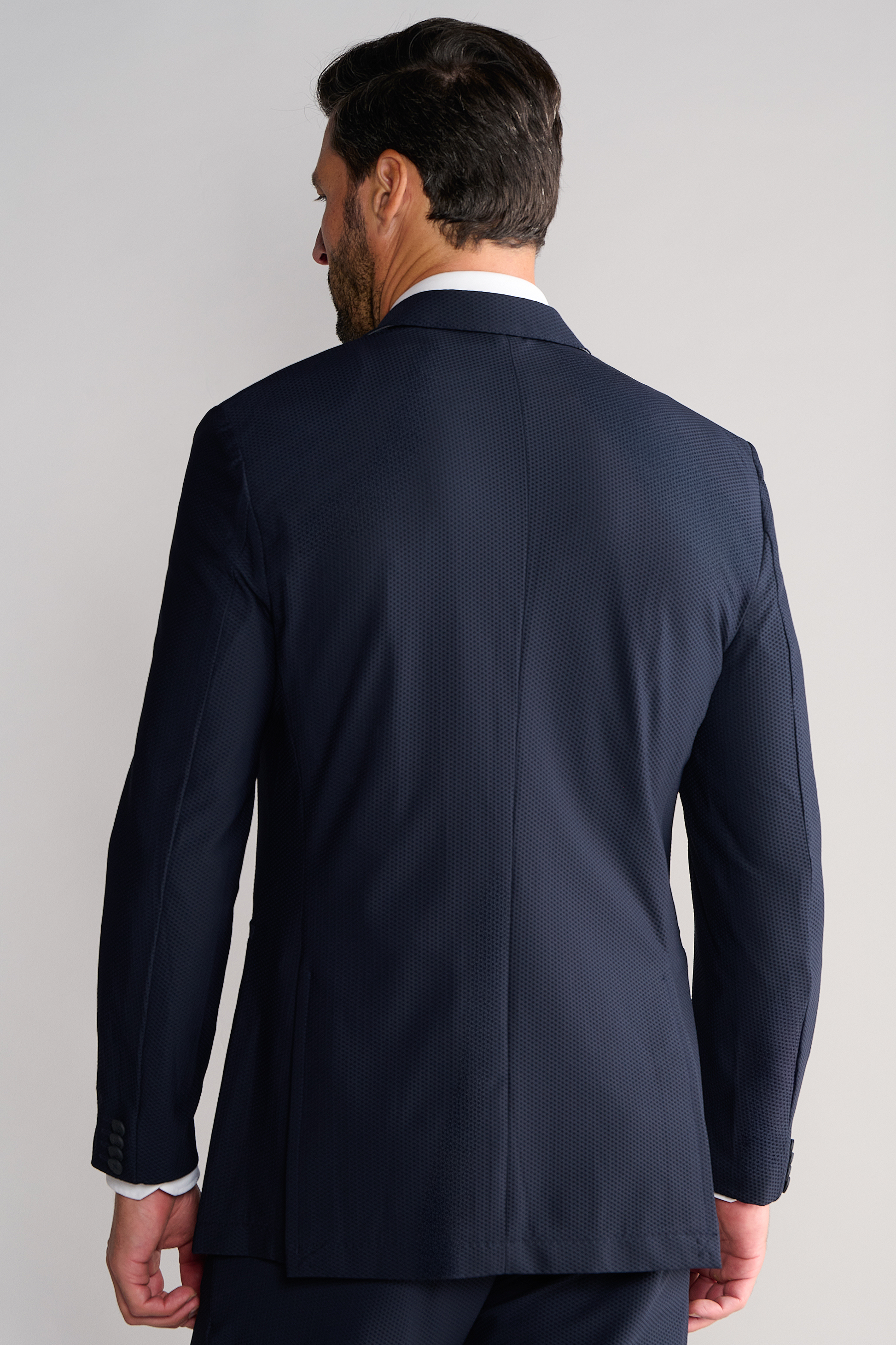 A man with dark hair and a short beard is seen from behind wearing Twillory’s AIR Blazer // Navy, a breathable, subtly textured suit jacket with buttoned cuffs, matched with navy pants against a light gray background.