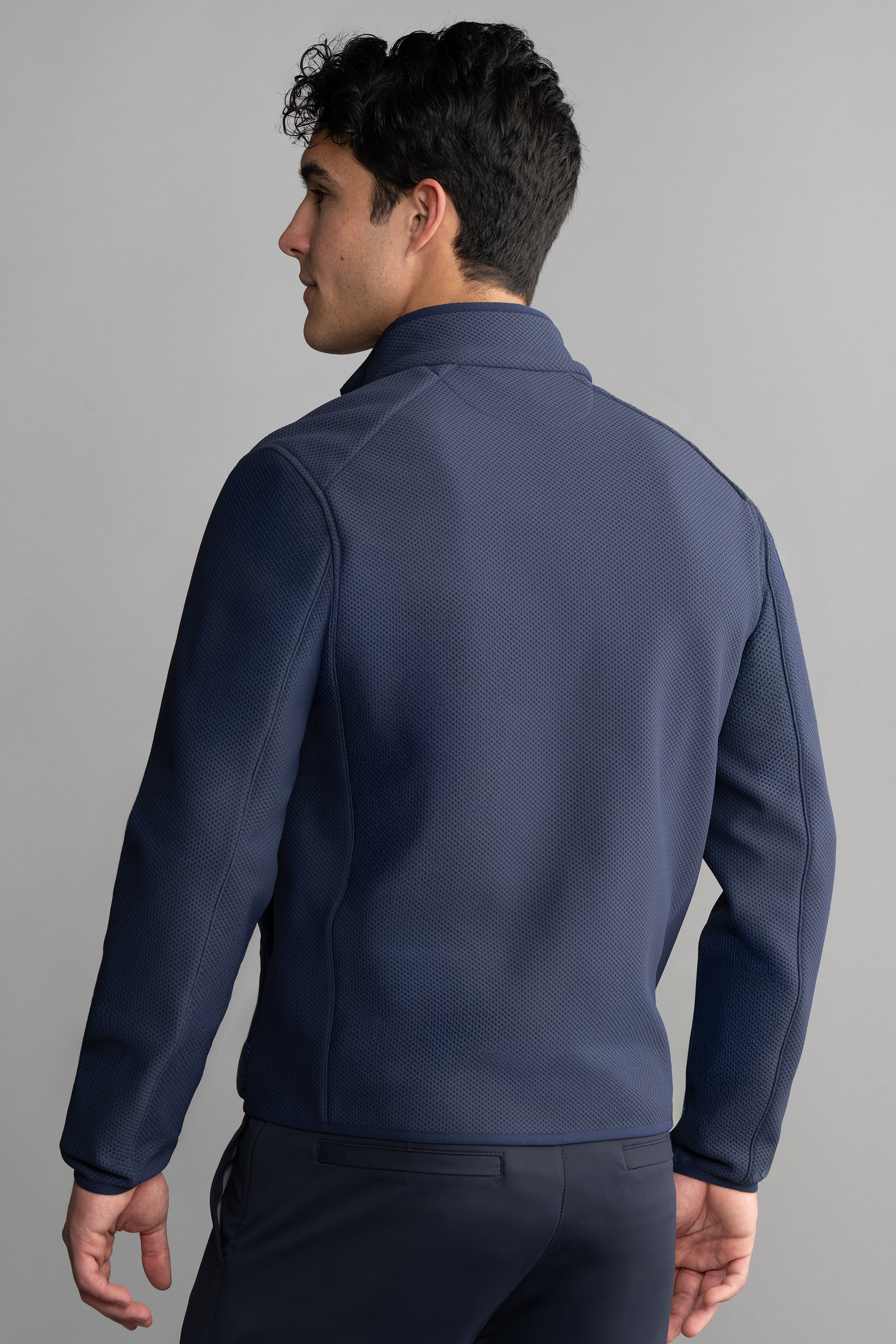 A man with short, dark, curly hair stands with his back to the camera, wearing Twillory’s Central Hybrid Jacket in navy. The background is plain light gray, and he is looking slightly to the left.