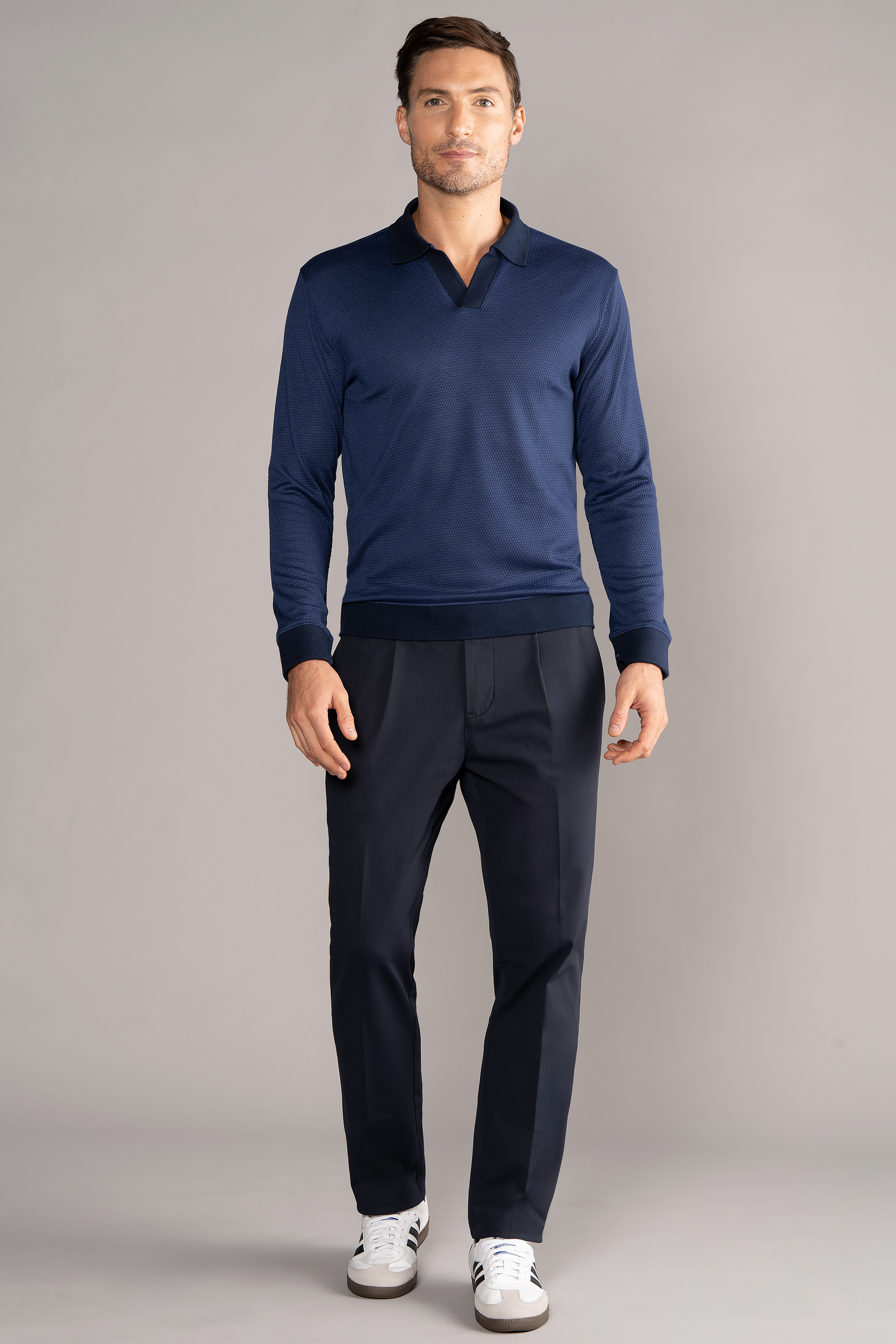 A man with short brown hair and a trimmed beard stands against a plain gray background, wearing the Twillory Long Sleeve Sweater Polo Solid in navy, dark navy dress pants, and white sneakers with black accents.