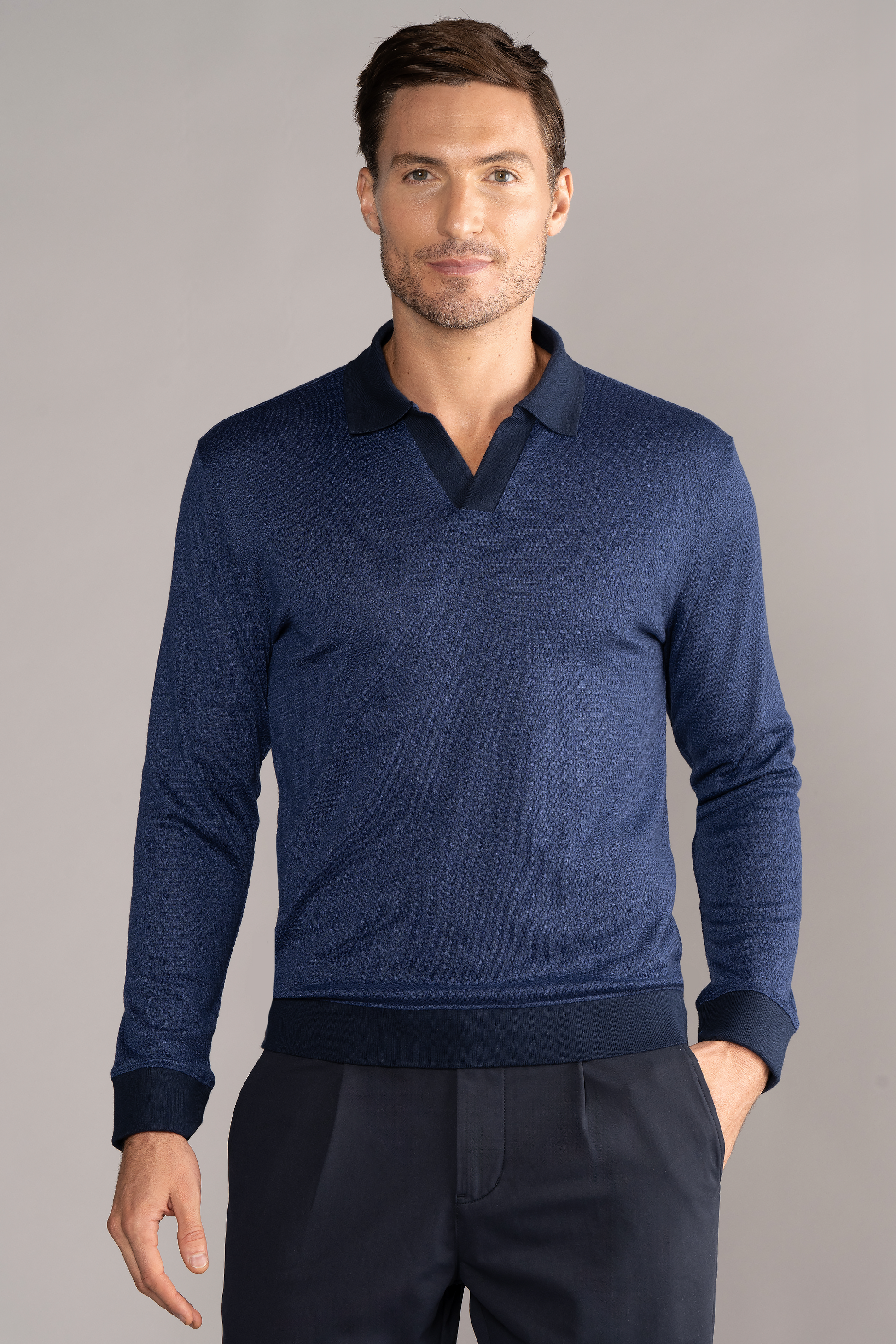 A man with short brown hair and a trimmed beard wears Twillory's Long Sleeve Sweater Polo Solid in navy, featuring dark cuffs and a V-neck Johnny collar, standing against a grey background with one hand in his trouser pocket.