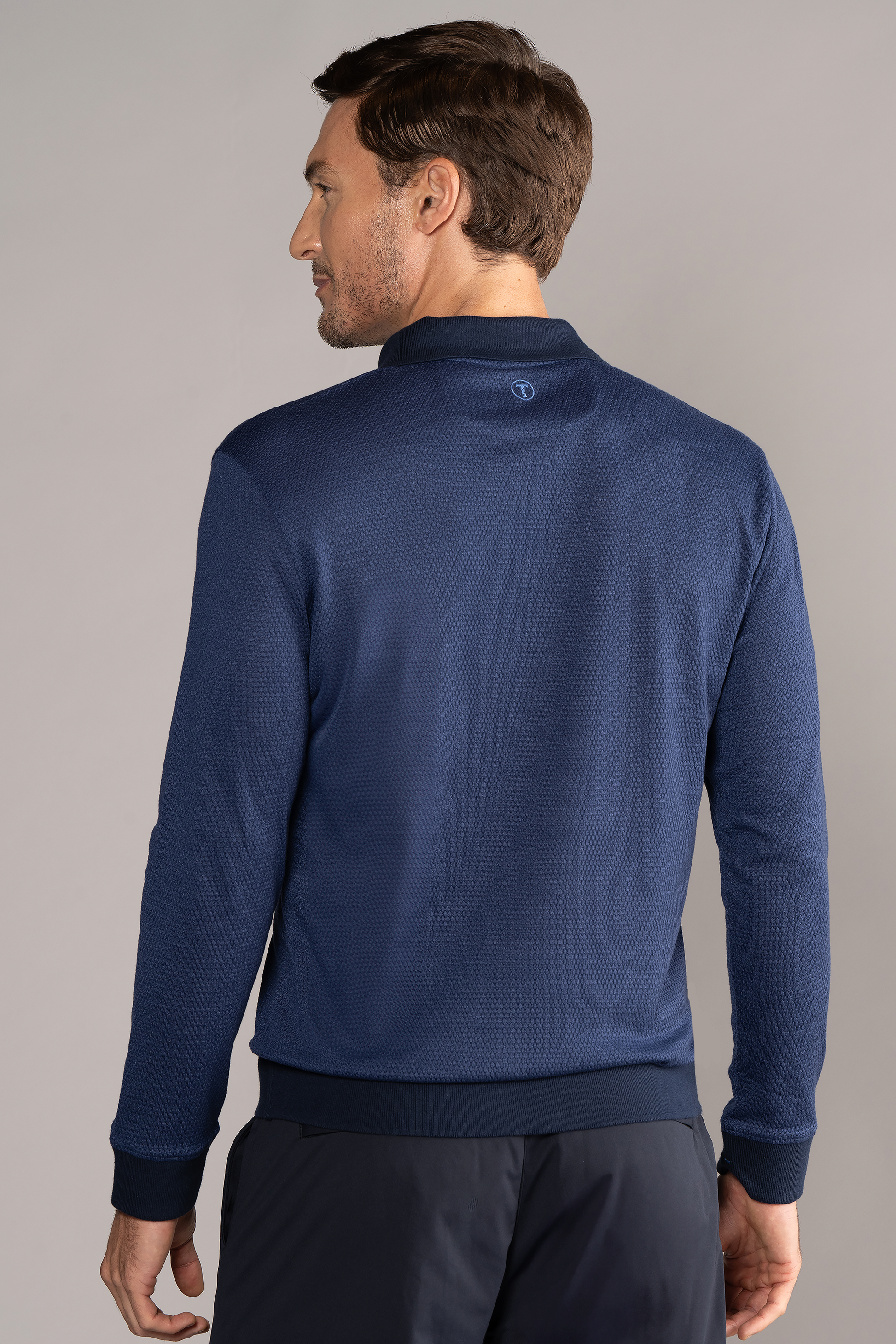 A man with short brown hair is seen from behind wearing the Twillory Long Sleeve Sweater Polo Solid in navy with matching pants, standing against a plain light gray background and looking slightly to his left.