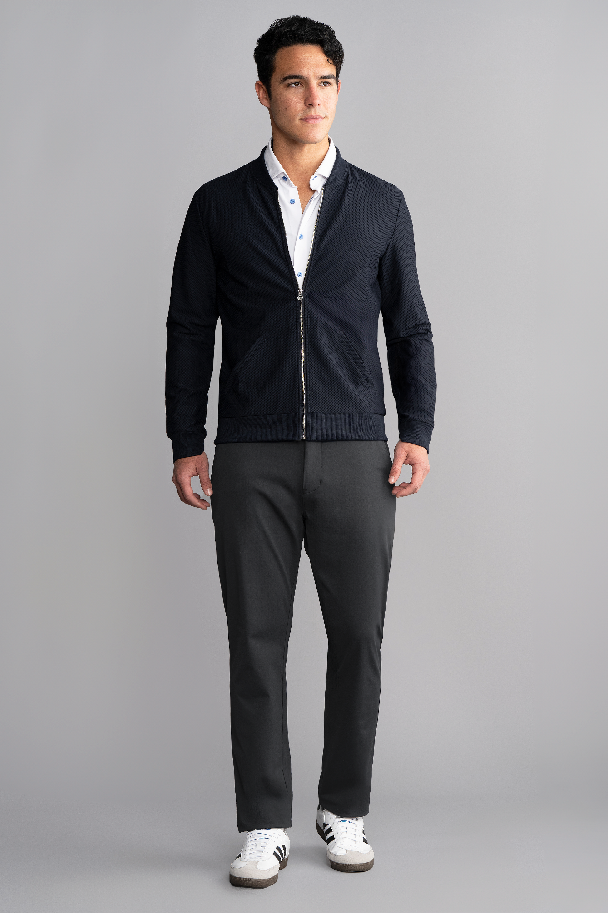 A man stands facing forward against a gray background, wearing the Twillory AIR Bomber in Navy over a white collared shirt, dark gray pants, and white sneakers with black stripes. His hair is short and dark; his expression is neutral.