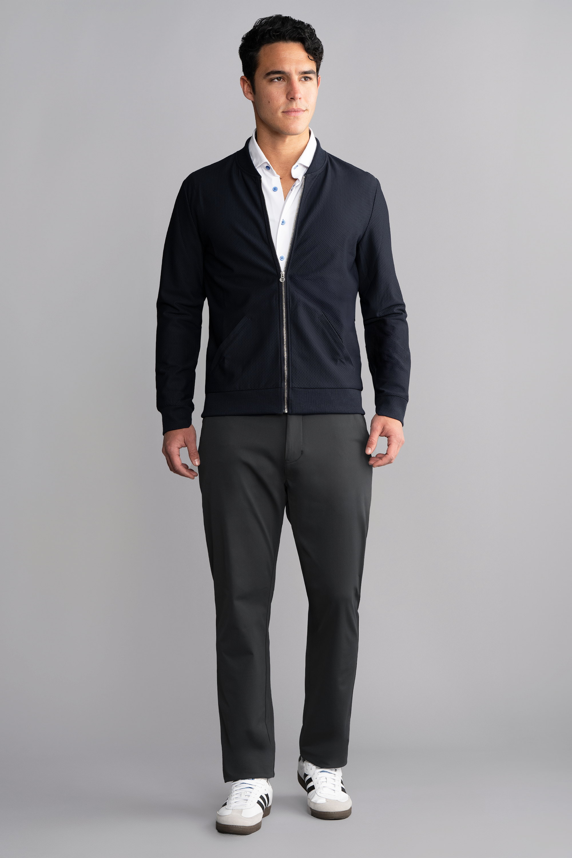 A man stands facing forward against a gray background, wearing the Twillory AIR Bomber in Navy over a white collared shirt, dark gray pants, and white sneakers with black stripes. His hair is short and dark; his expression is neutral.
