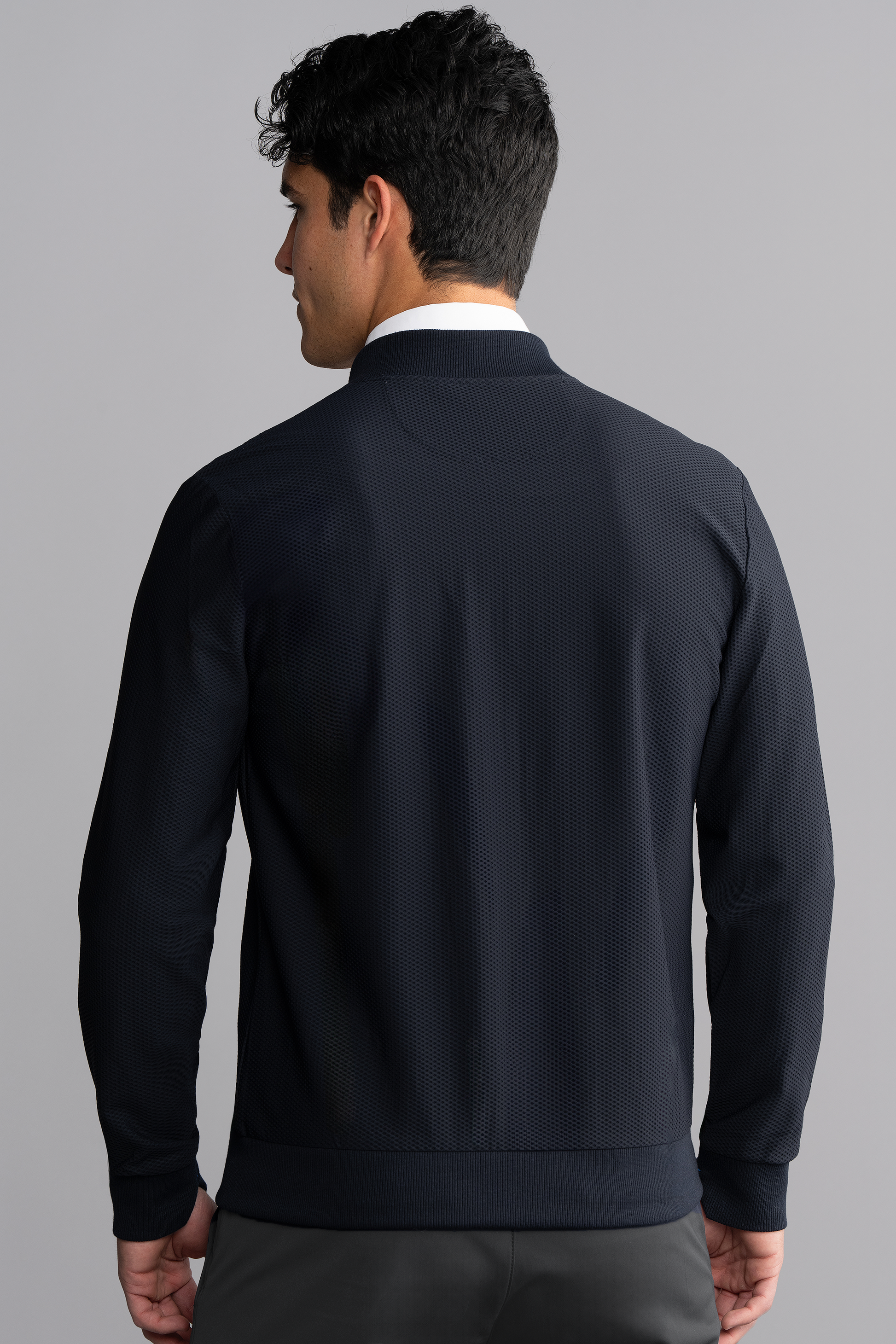 A man with short dark hair is seen from behind, wearing the Twillory AIR Bomber // Navy—crafted from 360° Airflow Fabric—over a white collared shirt, standing against a light gray background with his arms relaxed by his sides.