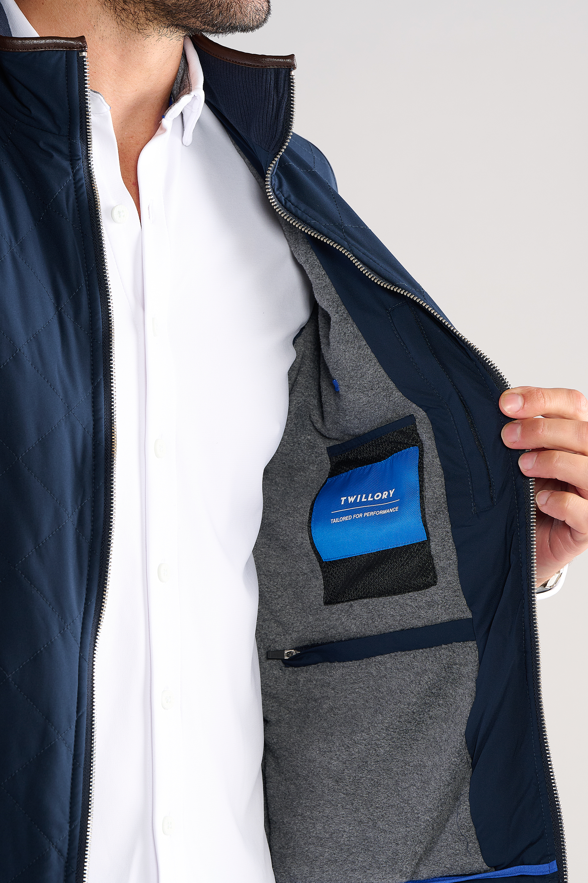 A man in a white shirt holds open a navy Performance Quilted Vest by Twillory, showing the grey lining with a blue pocket labeled "Twillory Designed for Performance" and a mesh pocket. Only his torso and lower face are visible.
