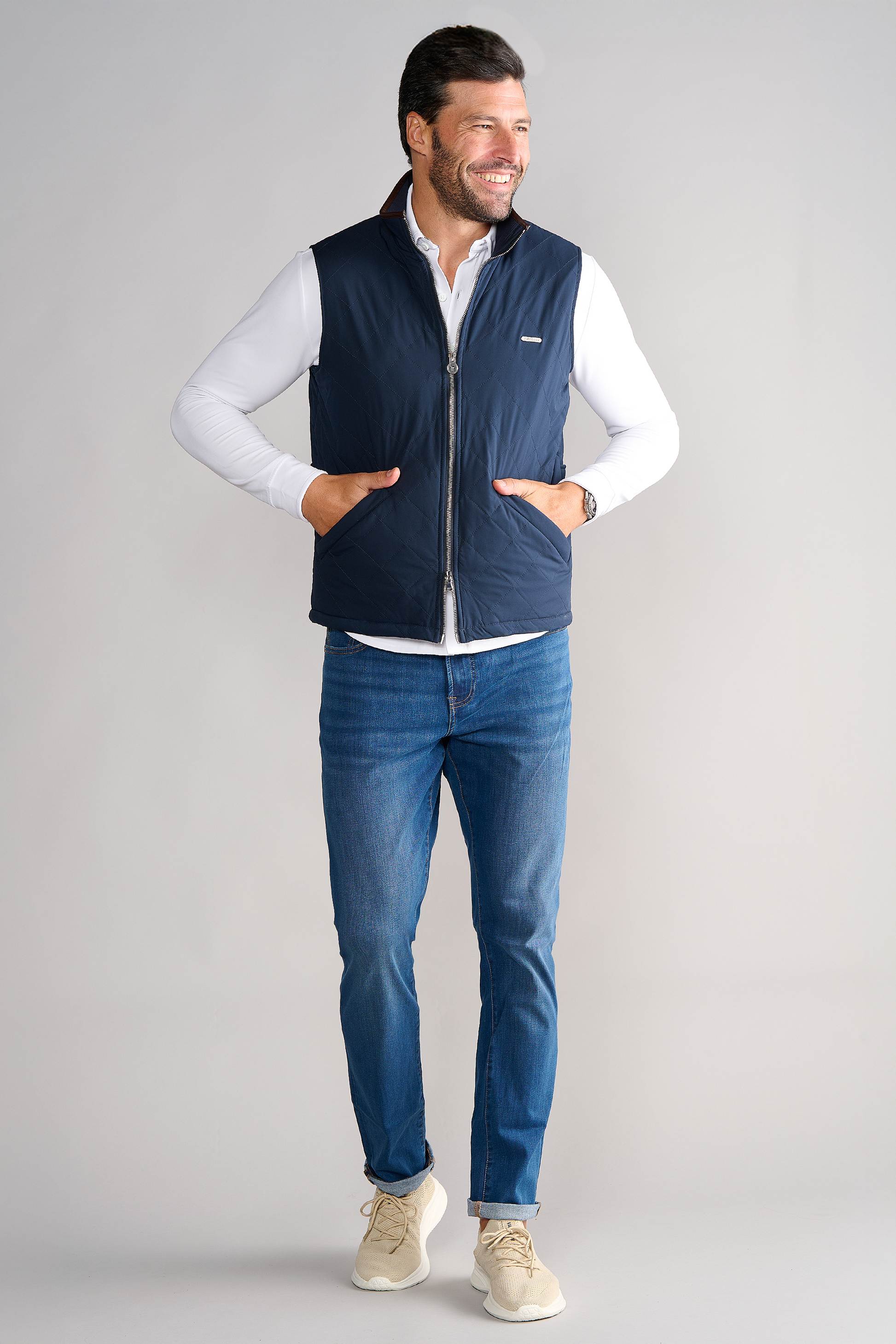 A smiling man with short dark hair and a trimmed beard stands against a light gray background, hands in pockets. He wears the Twillory Performance Quilted Vest over a white long-sleeve shirt, blue jeans, and beige sneakers.