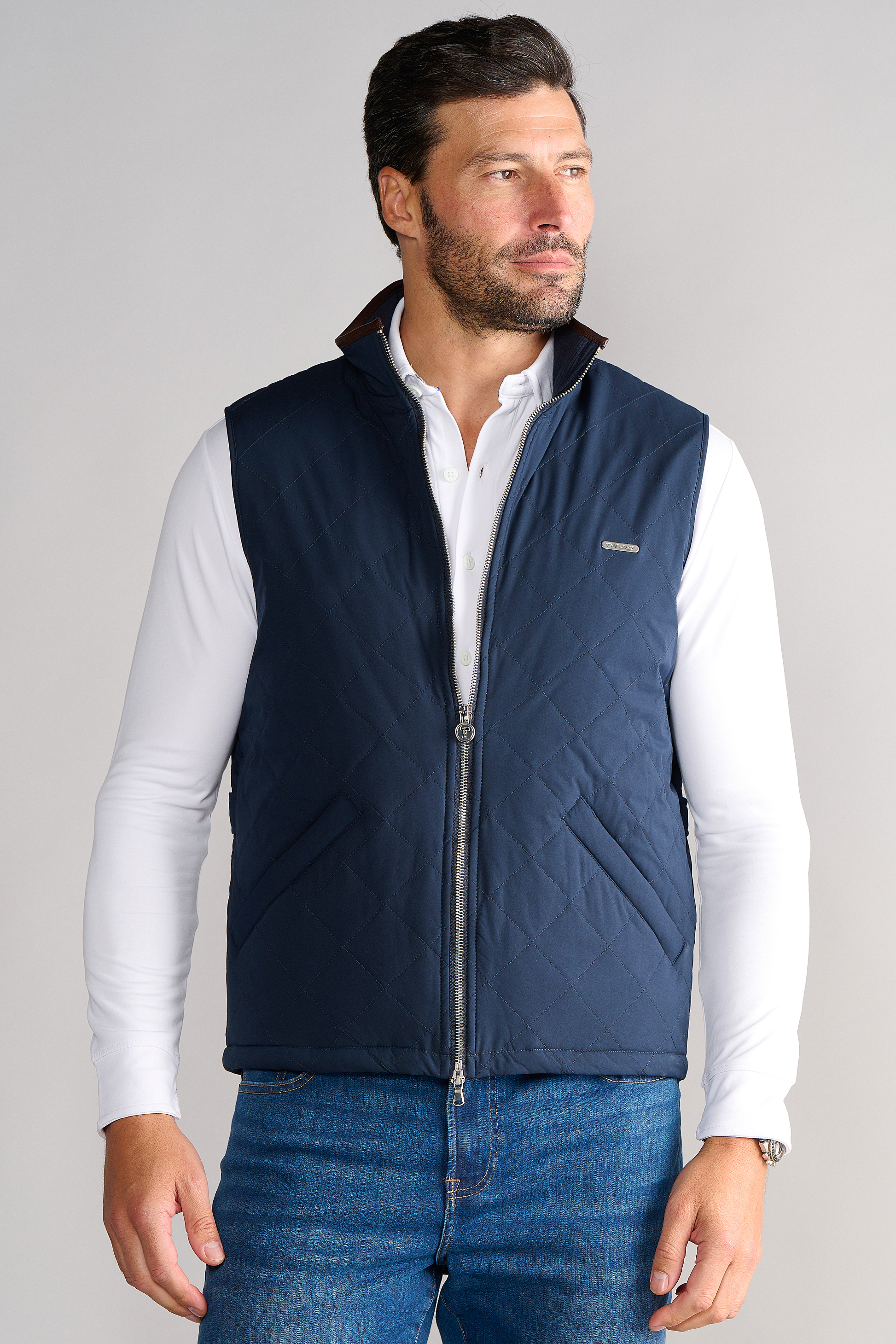 A dark-haired, bearded man wears a navy blue Twillory Performance Quilted Vest over a white button-down and blue jeans, standing against a plain light background and looking slightly to the left.