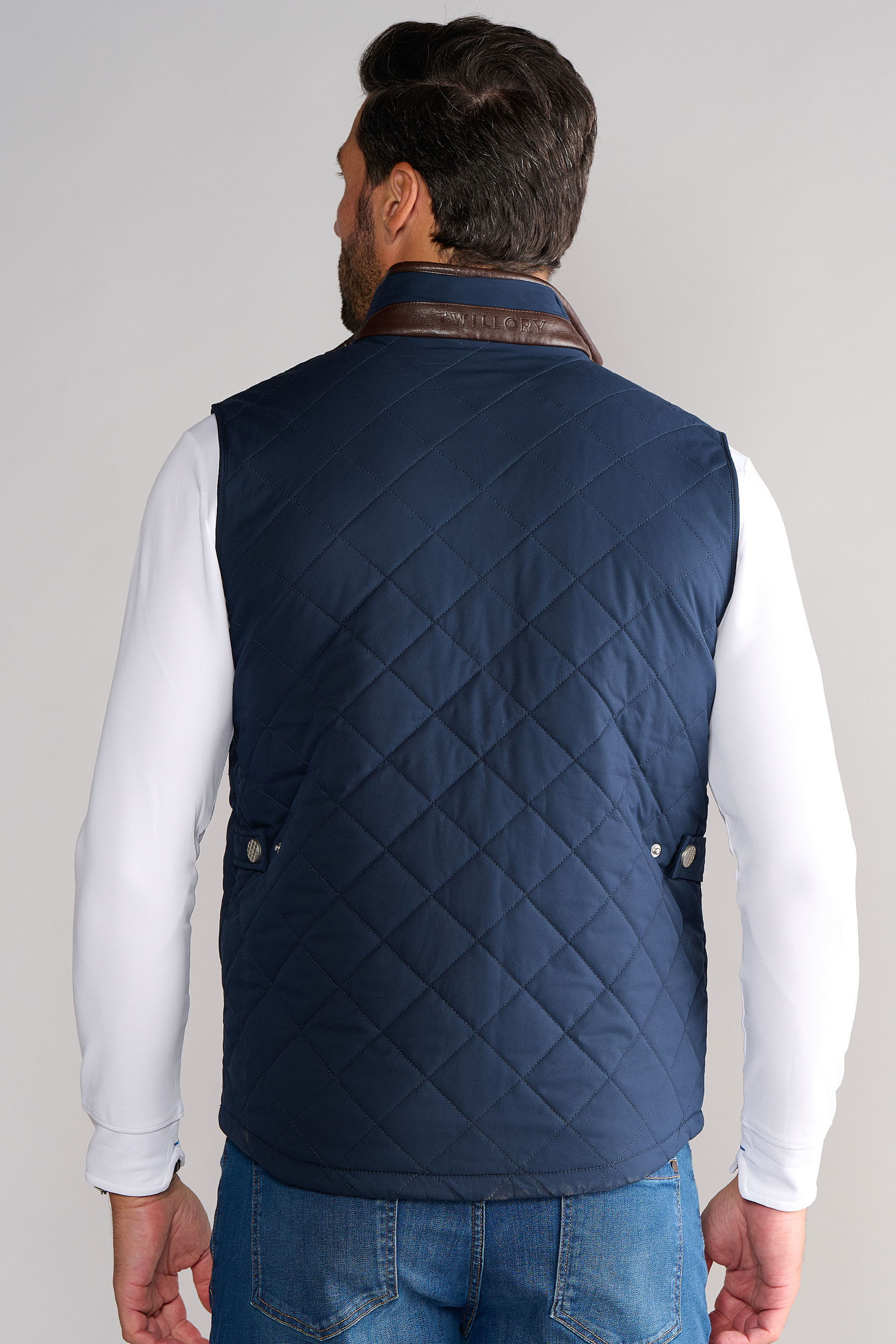 A man with short dark hair, seen from behind, wears a Twillory Performance Quilted Vest with a brown leather collar over a white long-sleeve shirt and blue jeans. He stands against a plain light gray background with his arms at his sides.